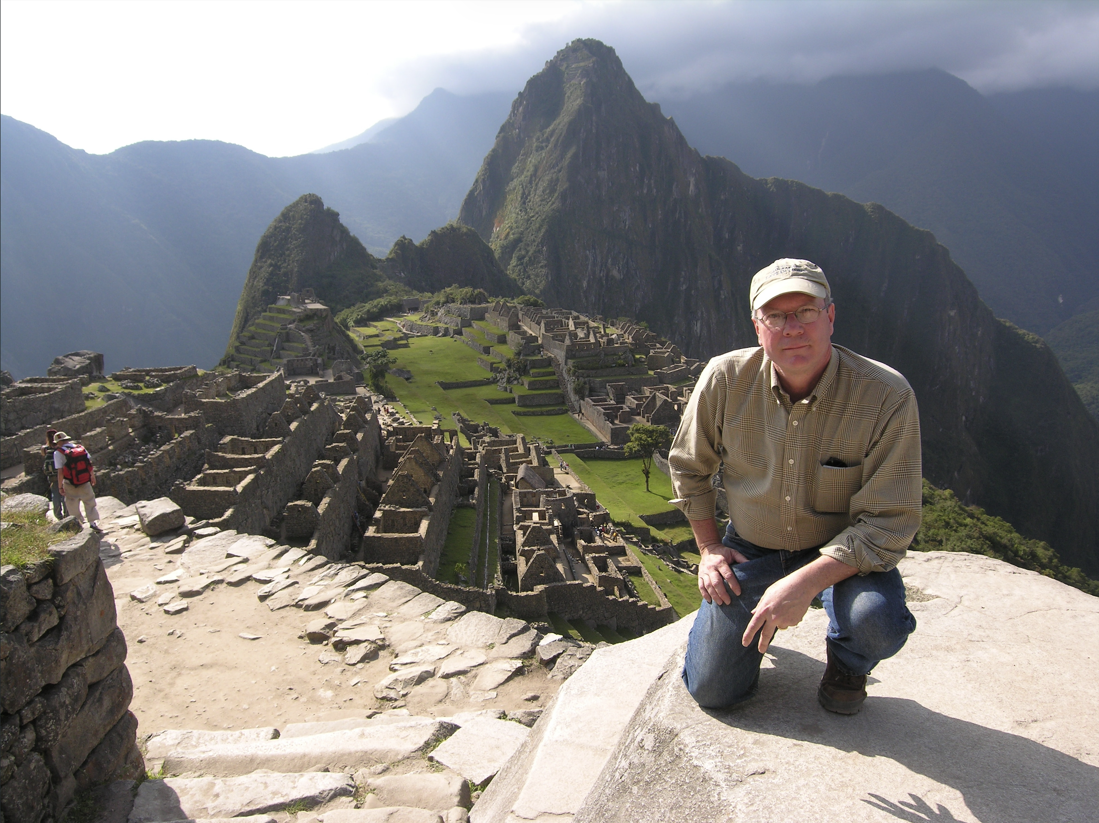 John Stubbs kneeling on a big rock in front of Machu Picchu, wearing a beige collared shirt, glasses, a baseball hat, and jeans 