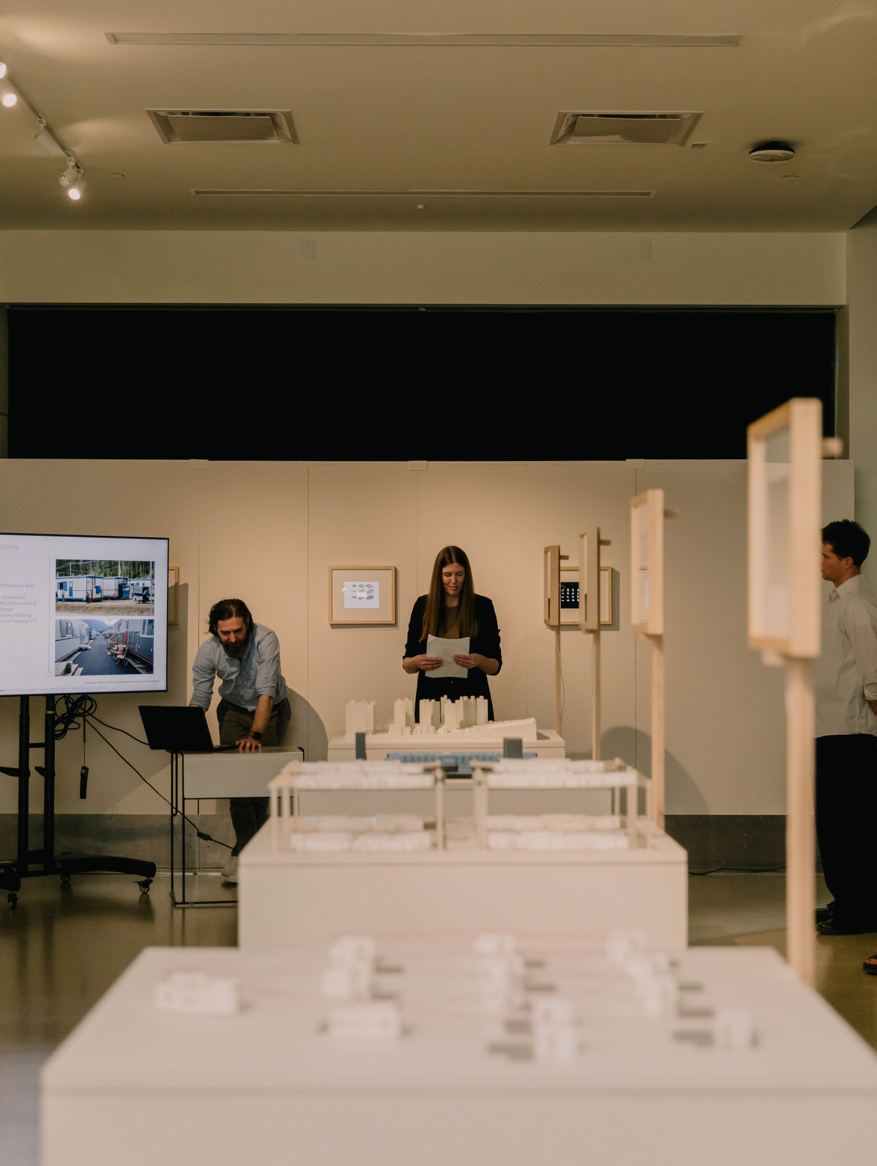 Small building models on tables in the middle of a gallery space with three standing people looking down at the models.