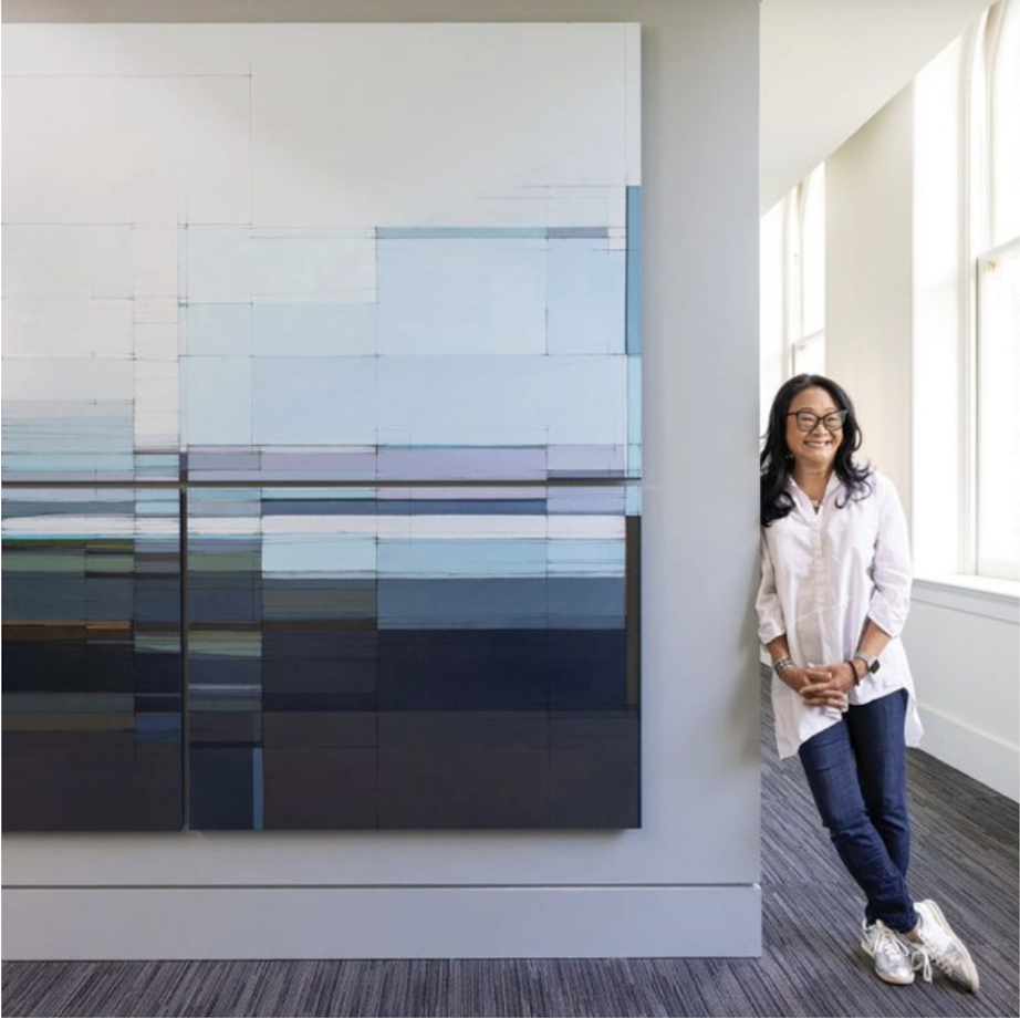 Tiffany Lin posed against a gallery wall with a large geometric blue, grey, and white painting, wearing a white shirt and jeans