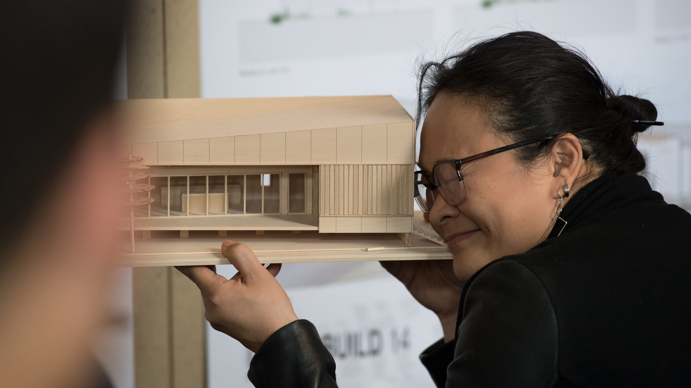 Photo of a woman wearing glasses holding up a wooden model and looking through the model with one eye closed.