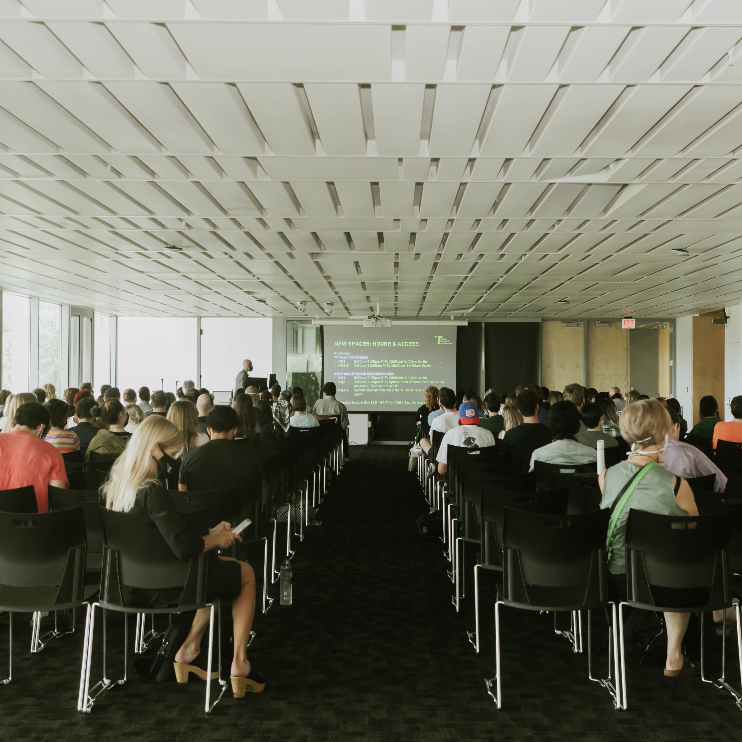 Picture taken from the back of a large auditorium, looking at the front of the room with rows of chairs and people and an aisle down the center