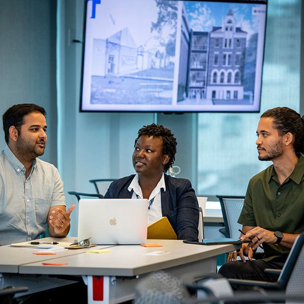 Three faculty sitting and talking at a table with a TV monitor screen in background showing architectural renderings