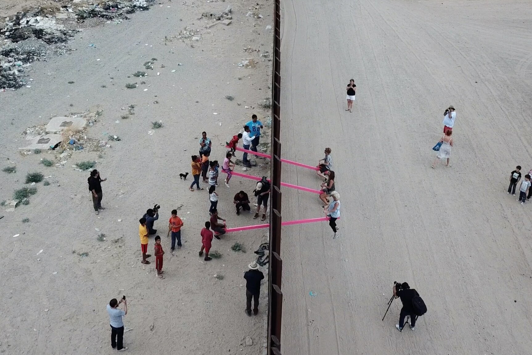 Aerial photo looking down on the U.S.-Mexico border wall as it runs vertically in the frame with people standing on either side of the wall and some people sitting on three vertical seesaws connected through the wall