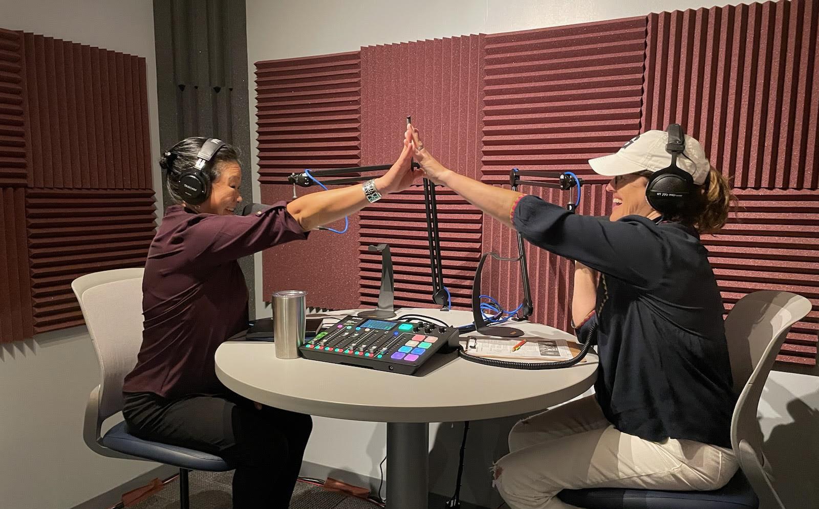 Two women, wearing headphones and speaking into large microphones, are sitting on either side of a round table inside a sound booth and are giving each other a high-five across the table