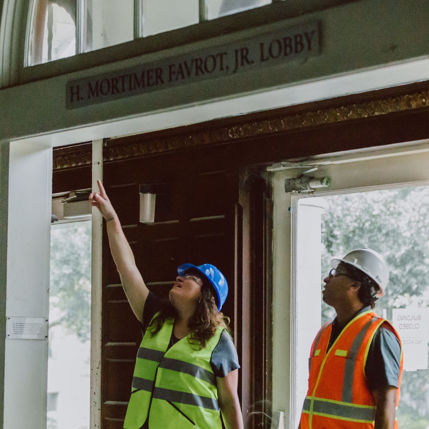 Two people wearing hard hats and safety vests stand inside the entrance of RMH lobby with one person pointing up to a beam 
