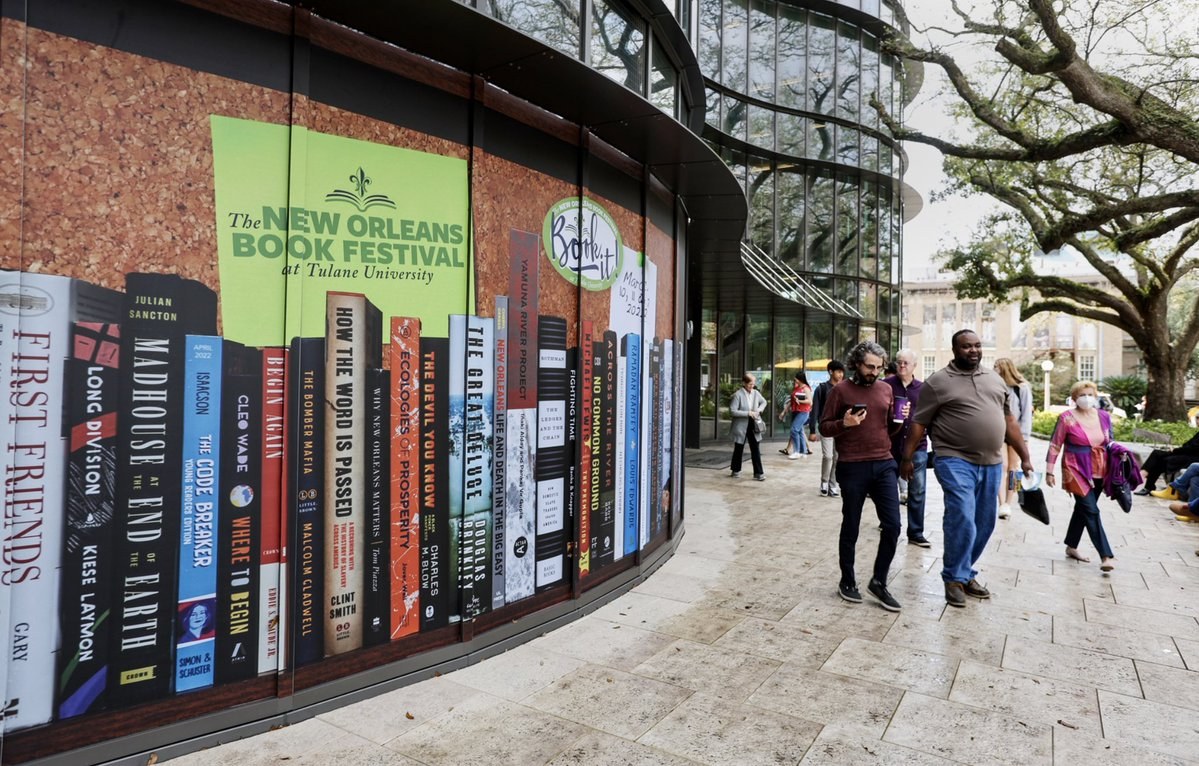A curved building exterior facade is wrapped with a large graphic decal of showing books lined up vertically while people walk next to the large sign on the right