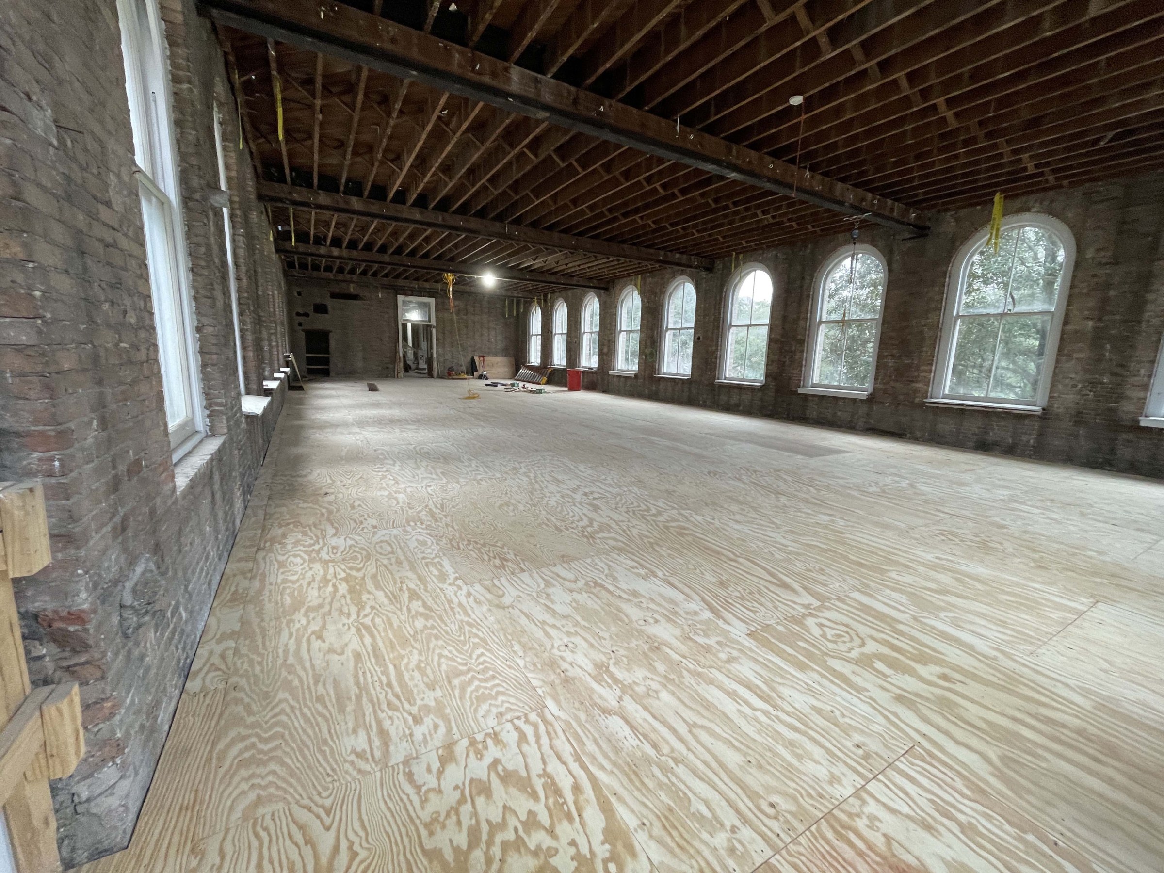 Interior of large room undergoing historic renovation with new wood floors and exposed ceiling beams high above.