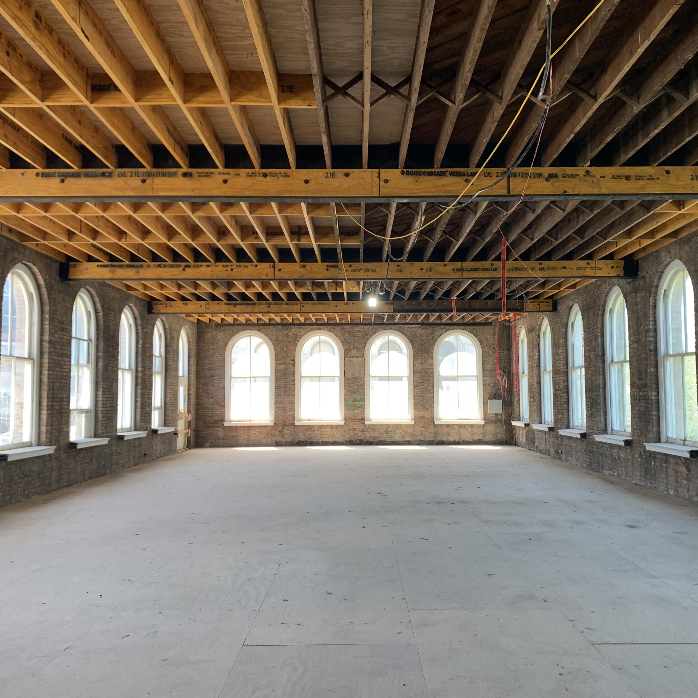 Interior view of a historic renovation inside a large hall with tall arched windows on lining the left, right, and back walls with exposed ceiling-floor beams above and a new sub-floor below.