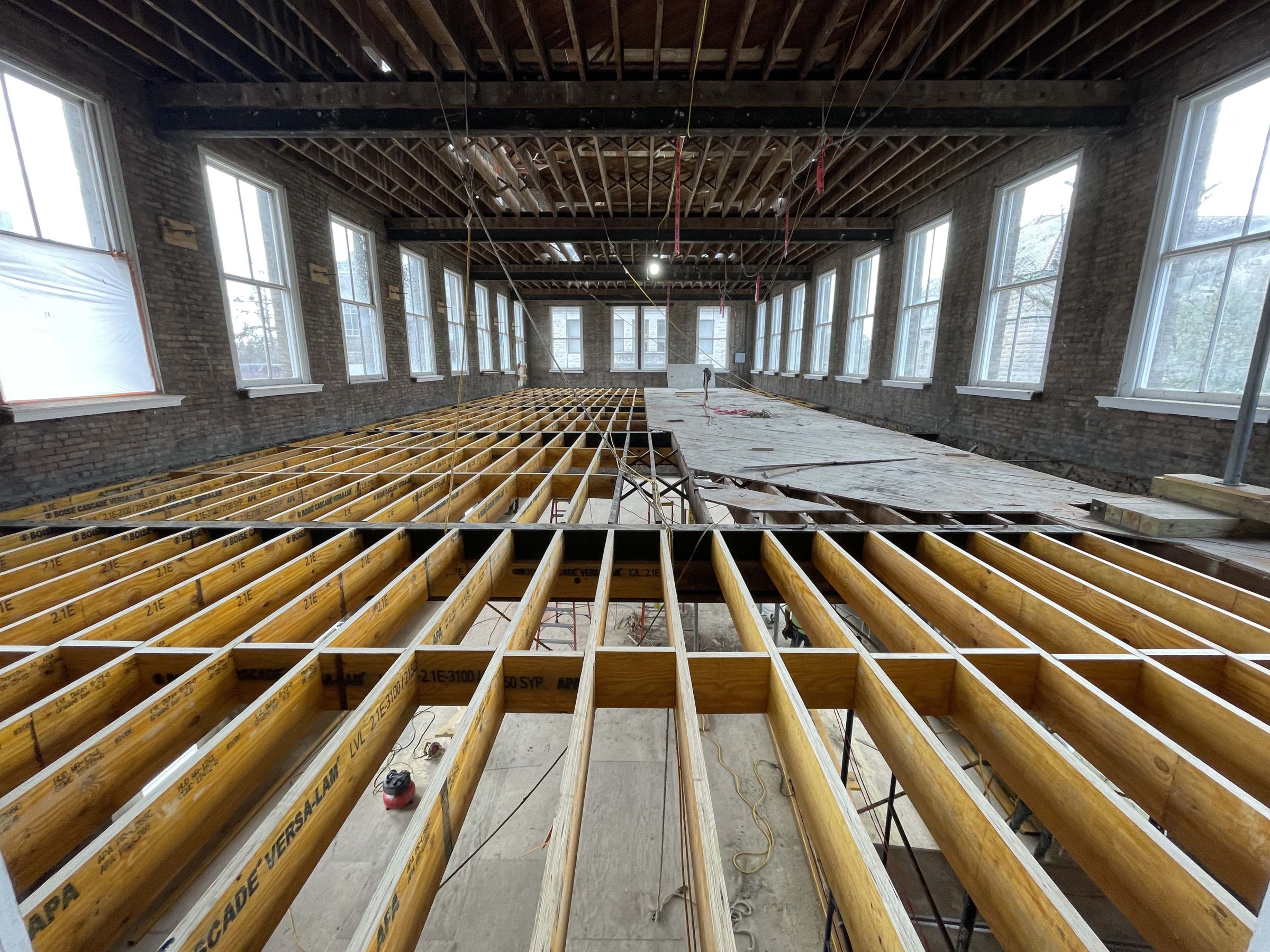 Interior photo of mid-renovation historic building with exposed beams for a new sub-floor running vertical in the picture and large windows on either side and back wall of the large room