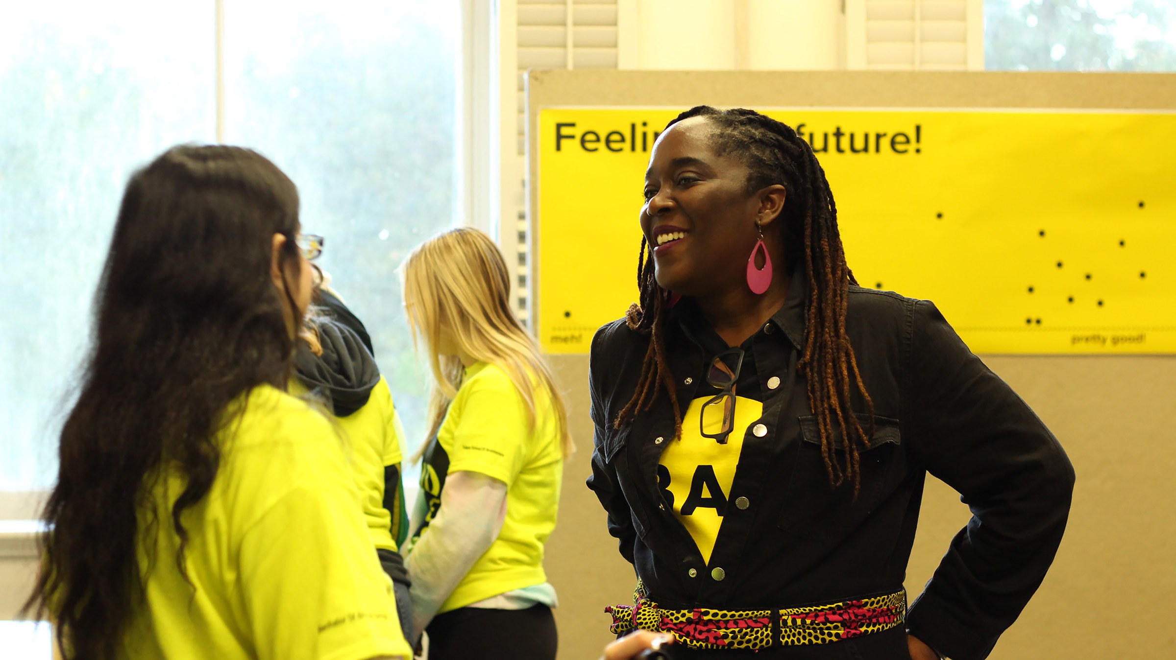 Photo of two people speaking to each other in front of bright yellow sign, with other people in the background