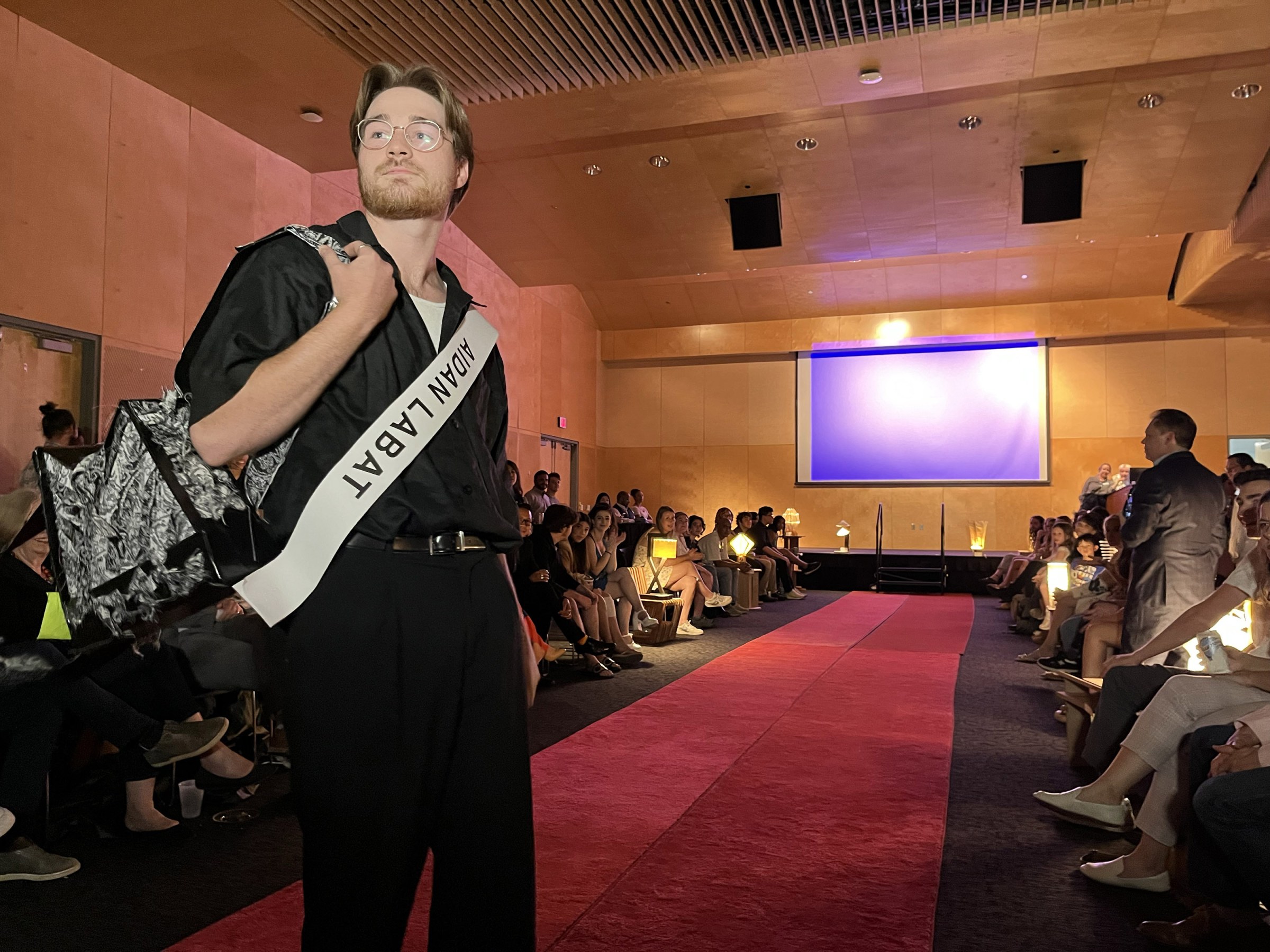 A model poses at the end of a carpeted catwalk to show a backpack design with people seated on both sides of the runway in the background