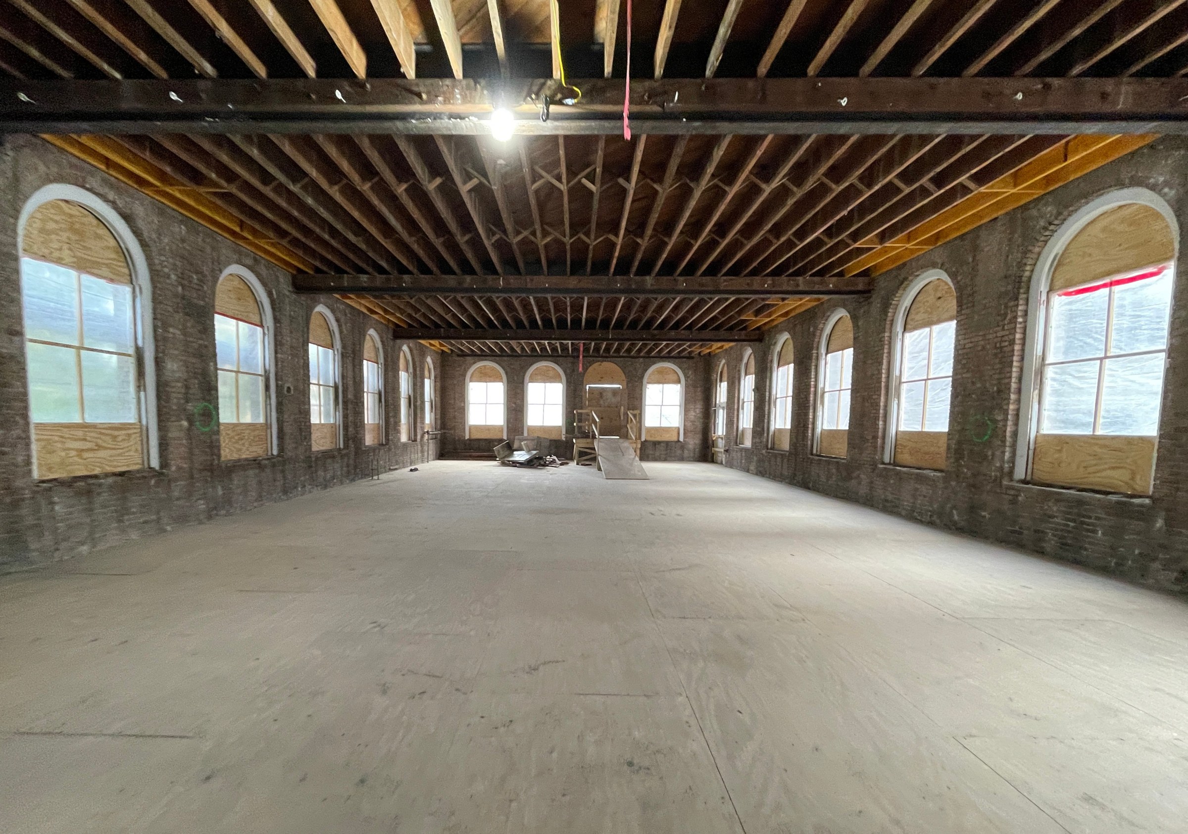 Interior of large room undergoing historic renovation with new wood floors and exposed ceiling beams high above.