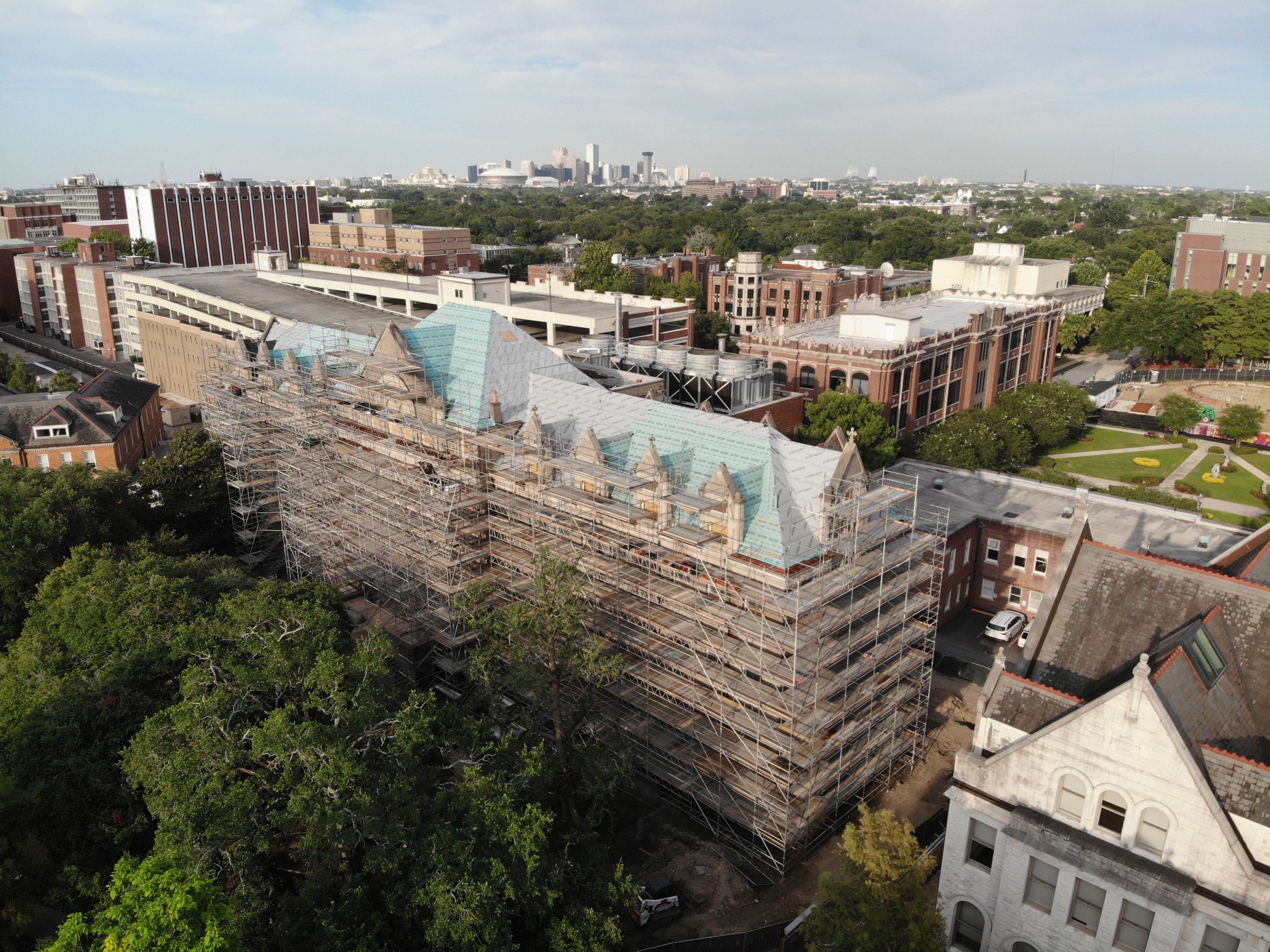 aerial view of southwest corner of Richardson Memorial Hall with scaffolding surrounding the building, trees in the foreground and Loyola's campus buildings in the background and New Orleans skyline in the distant background.