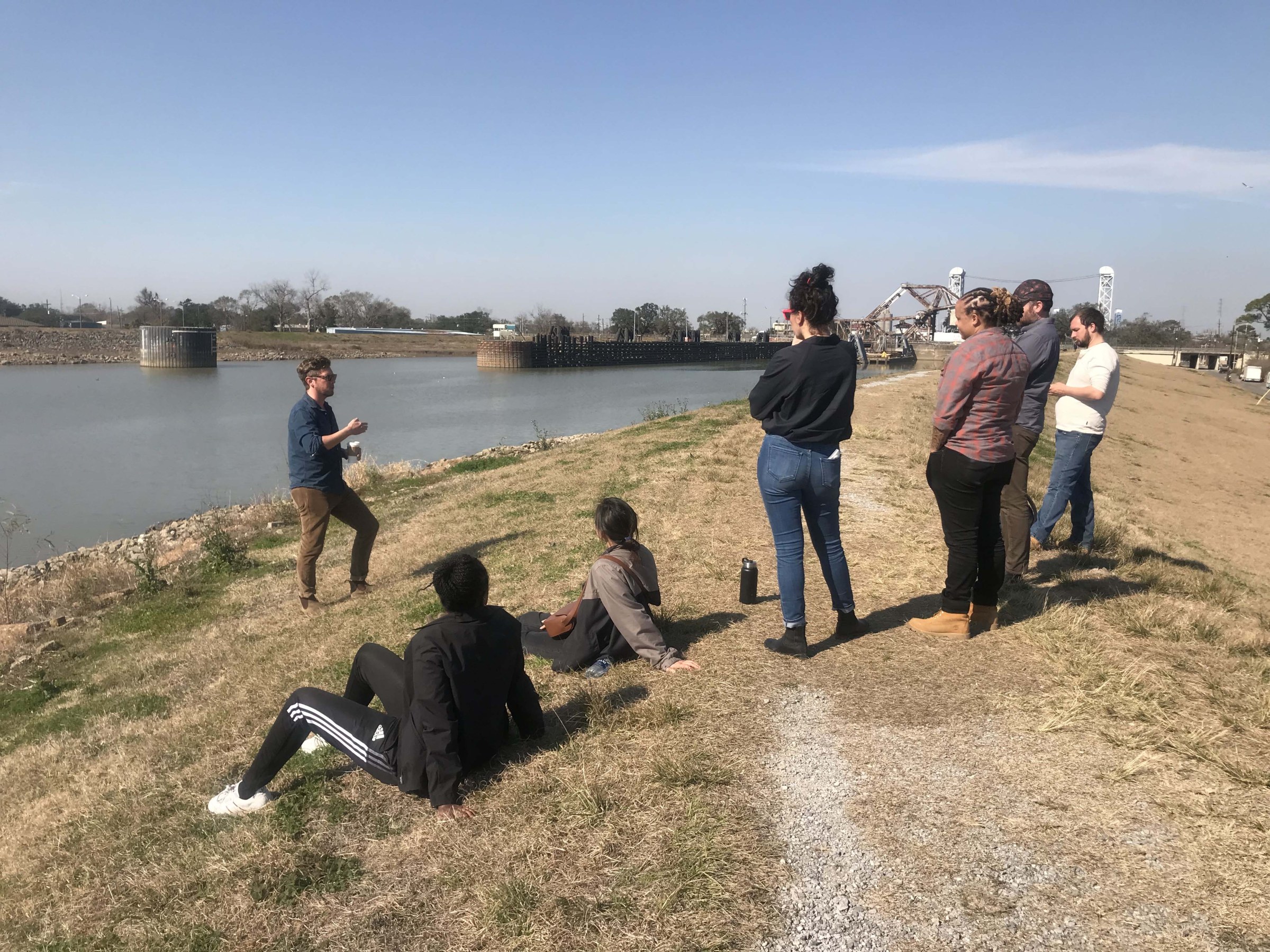 A small group of people sit and stand while talking on an earthen levee next to an industrial canal with an bridge in the background.