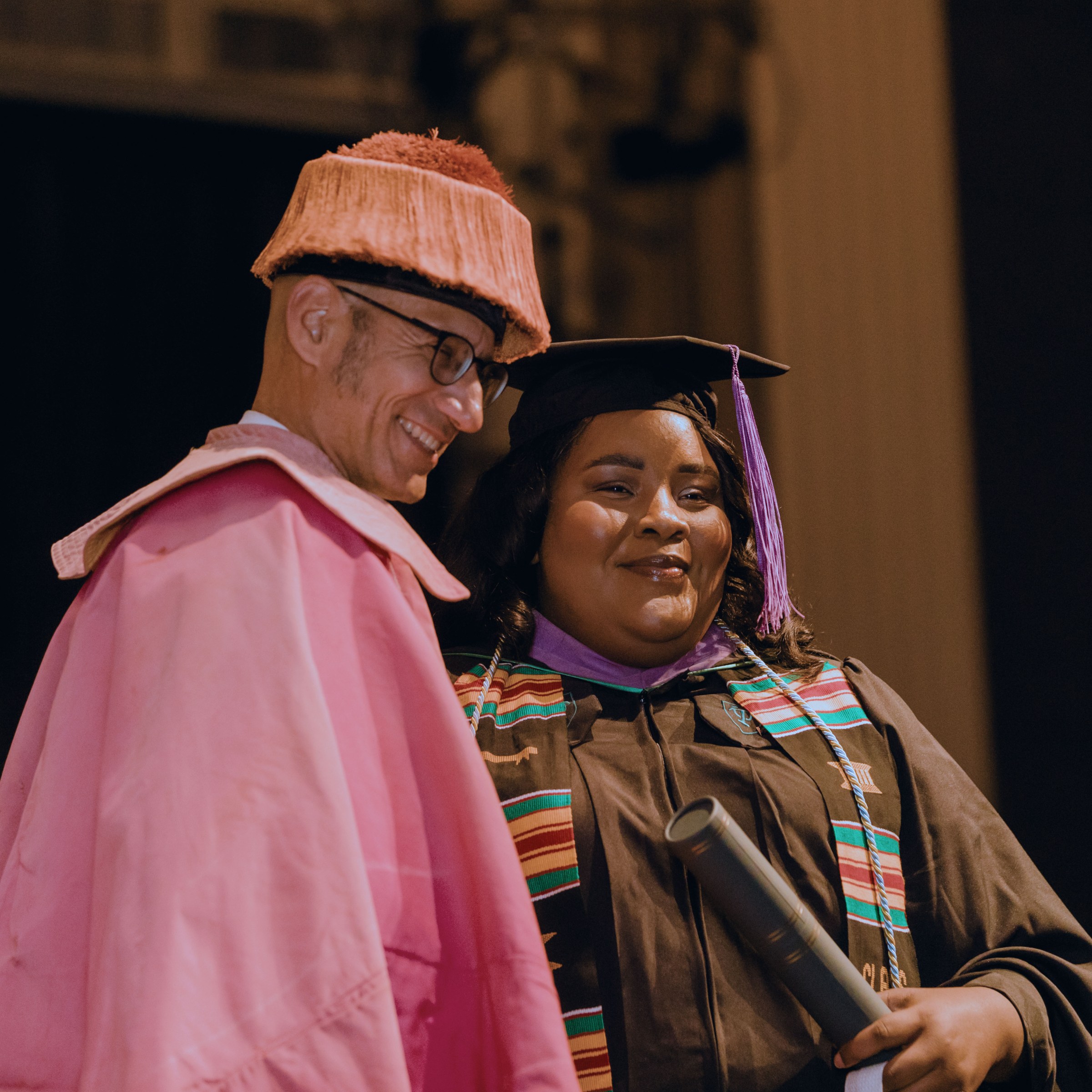 A professor and graduate in commencement regalia stand next to each other and pose for a photo at the ceremony, looking off camera.