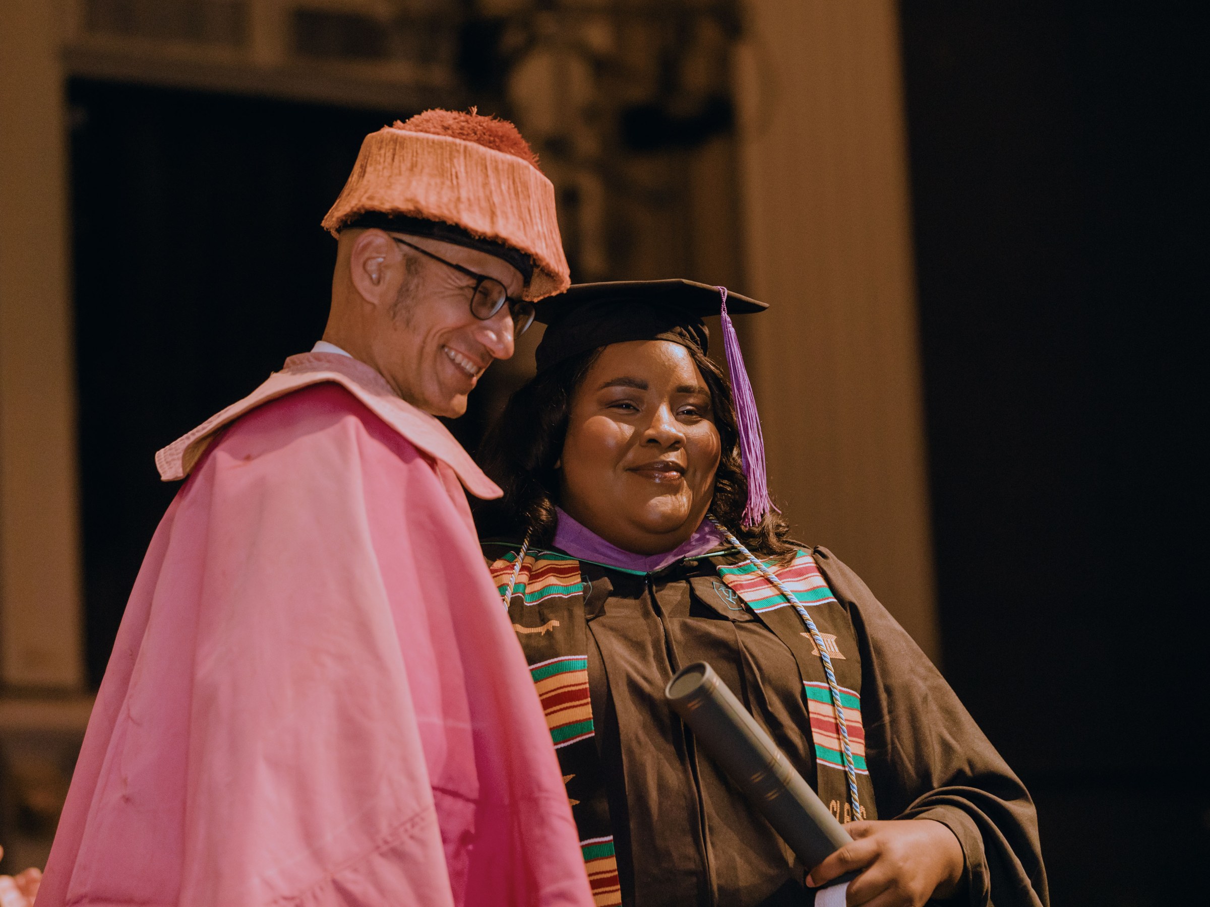 A professor and graduate in commencement regalia stand next to each other and pose for a photo at the ceremony, looking off camera.