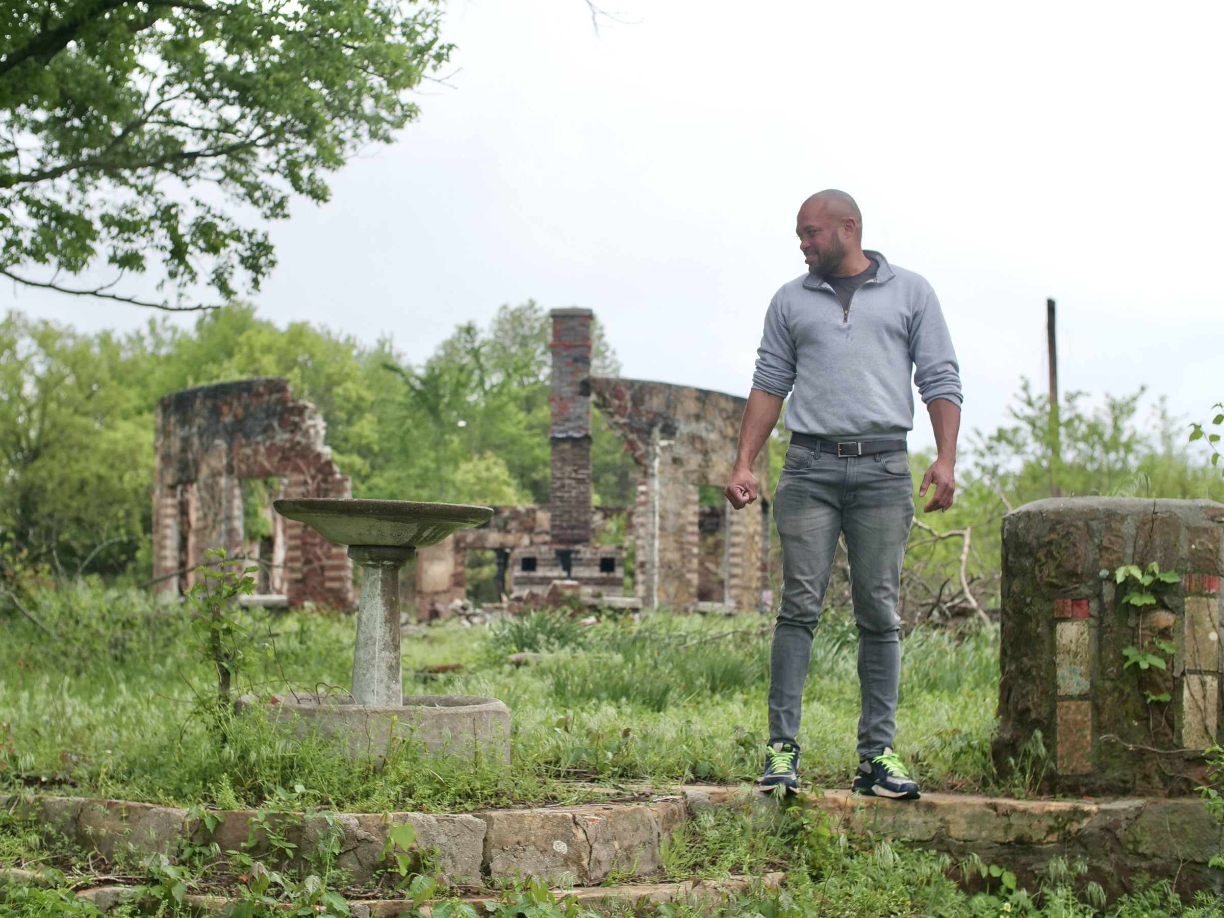 A man stands among ruins of a building or outdoor space with vegetation taking over much of the area and a small fountain to the right of him.