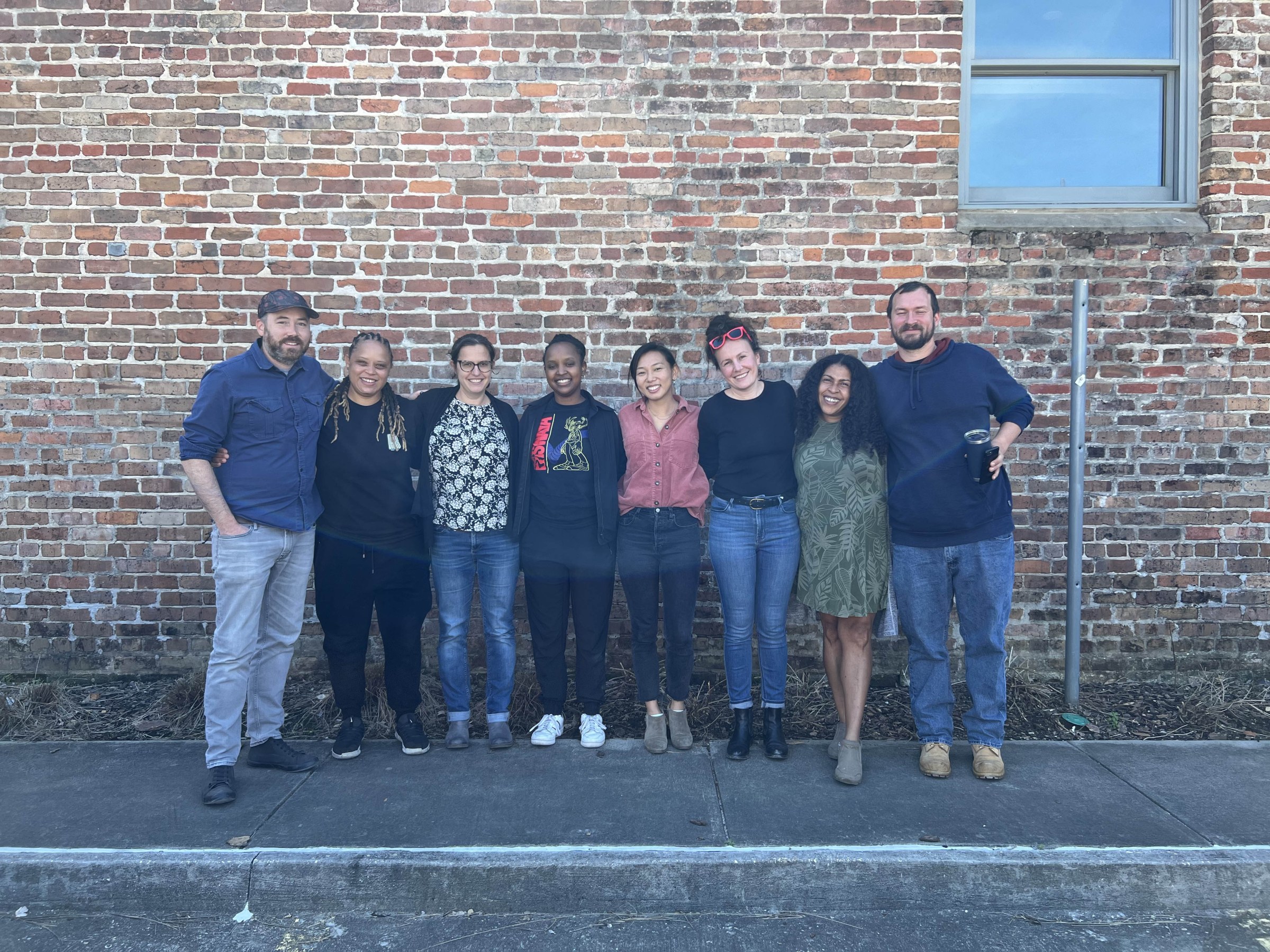 The Plot of Land team of eight people stand for a group photo, arms around each others shoulders, standing in front of a brick background.