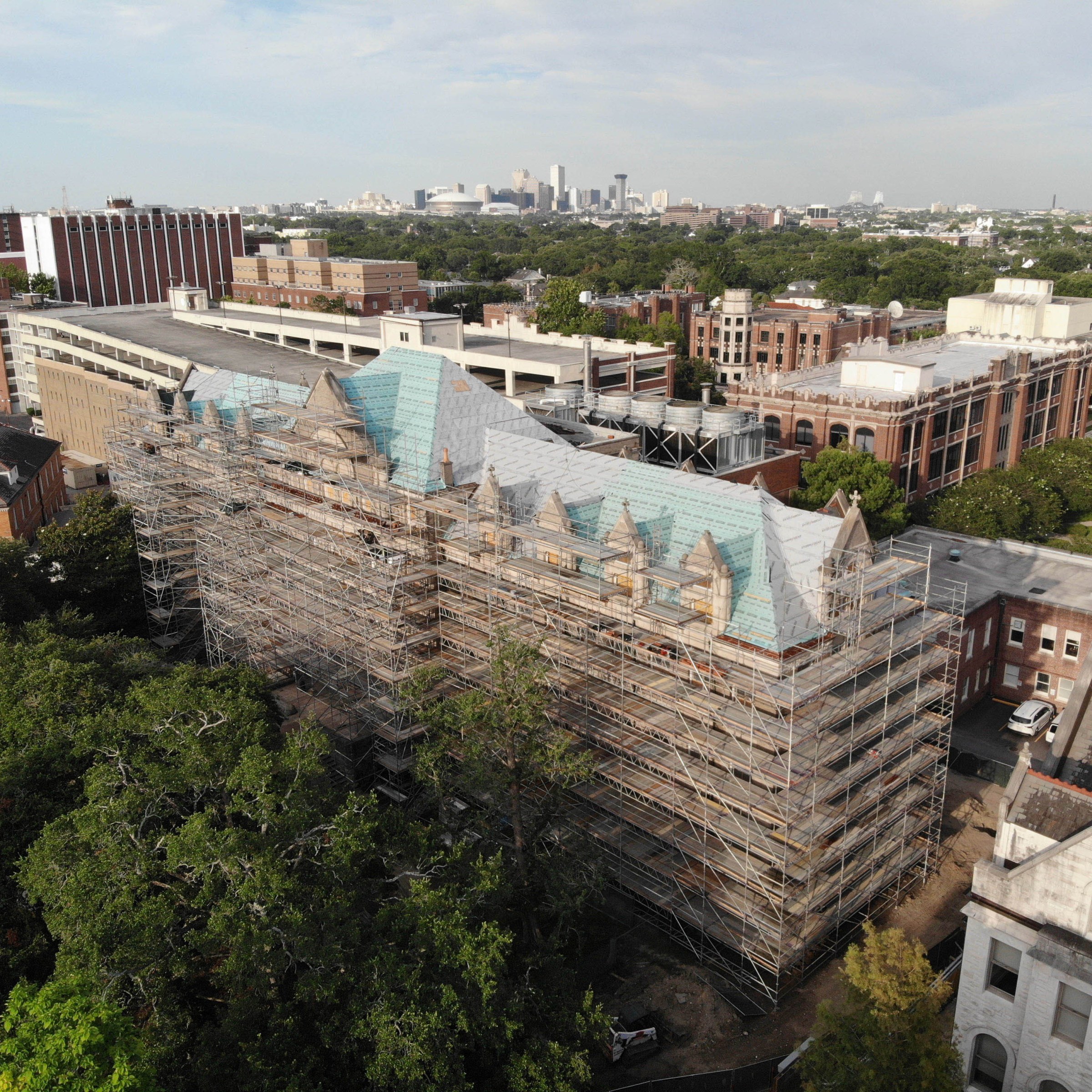 aerial view of southwest corner of Richardson Memorial Hall with scaffolding surrounding the building, trees in the foreground, Loyola's campus buildings in the background and New Orleans skyline in the distant background.