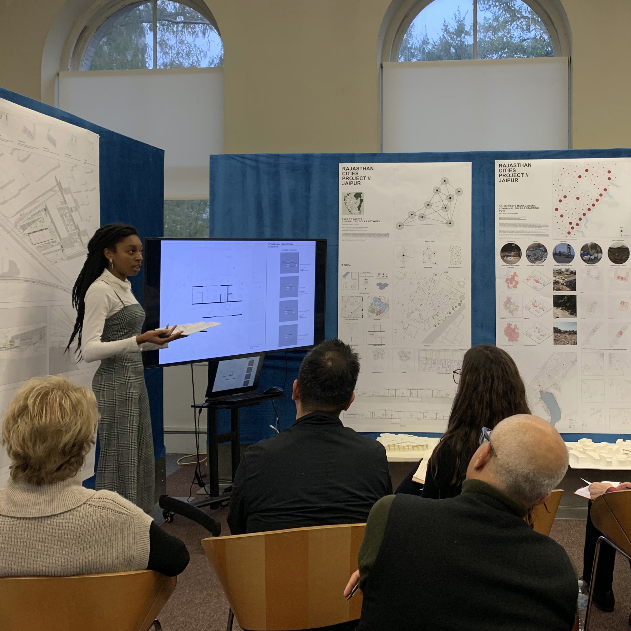 A student stands to present digital slides on a large screen in front of a panel of guest critics.