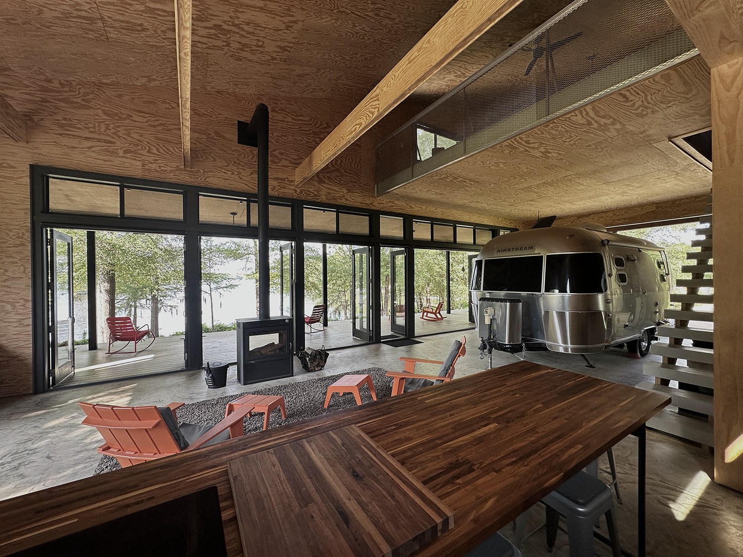 Interior of a open floor plan living space with a wall of windows leading to a porch in the background and on the right a roll-up door/wall that is open and an airstream trailer is pulled into the living space.