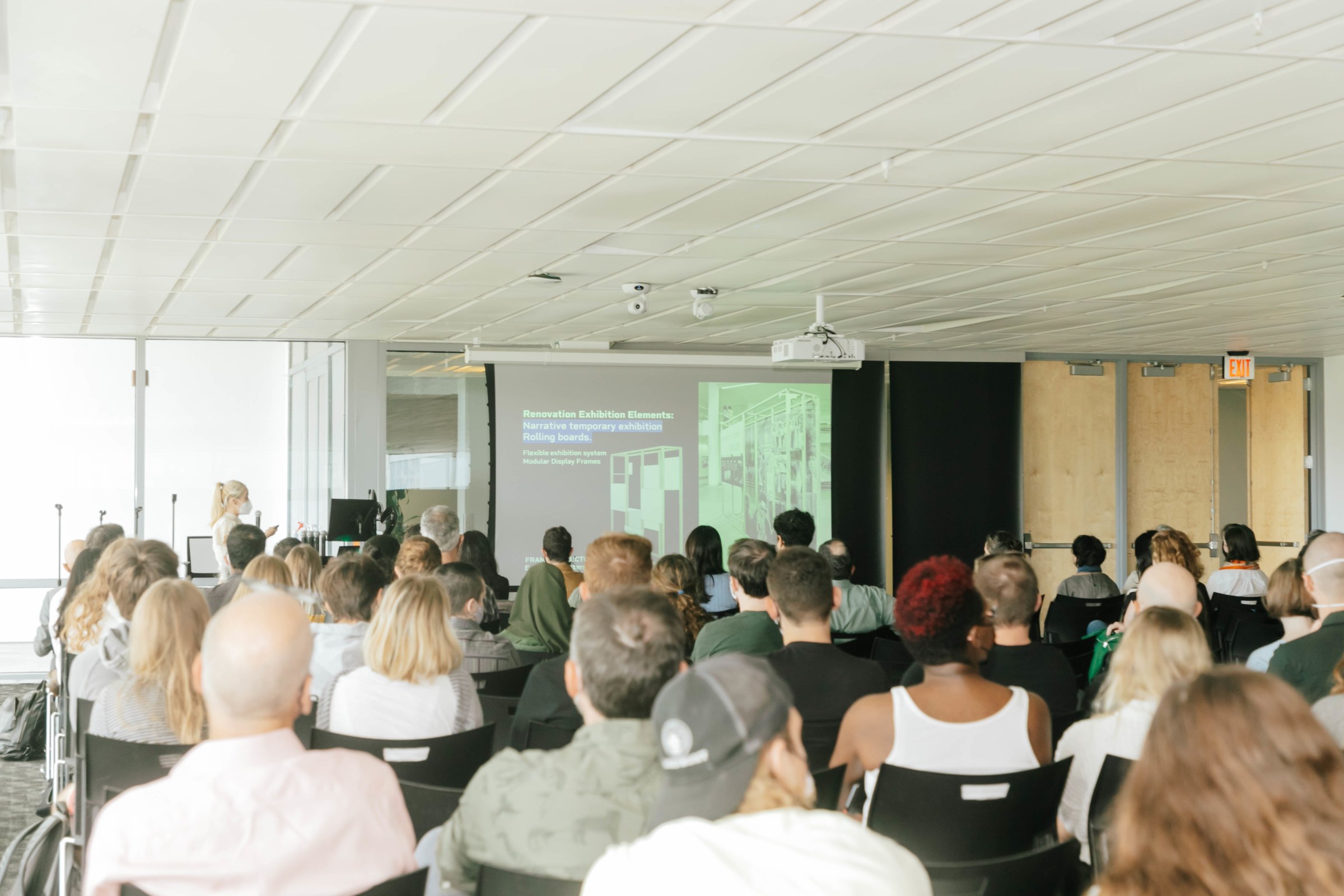 Photo from the back of a lecture hall room, looking toward the front of the room, over the heads of many people sitting in chairs looking at slides on a projector screen.