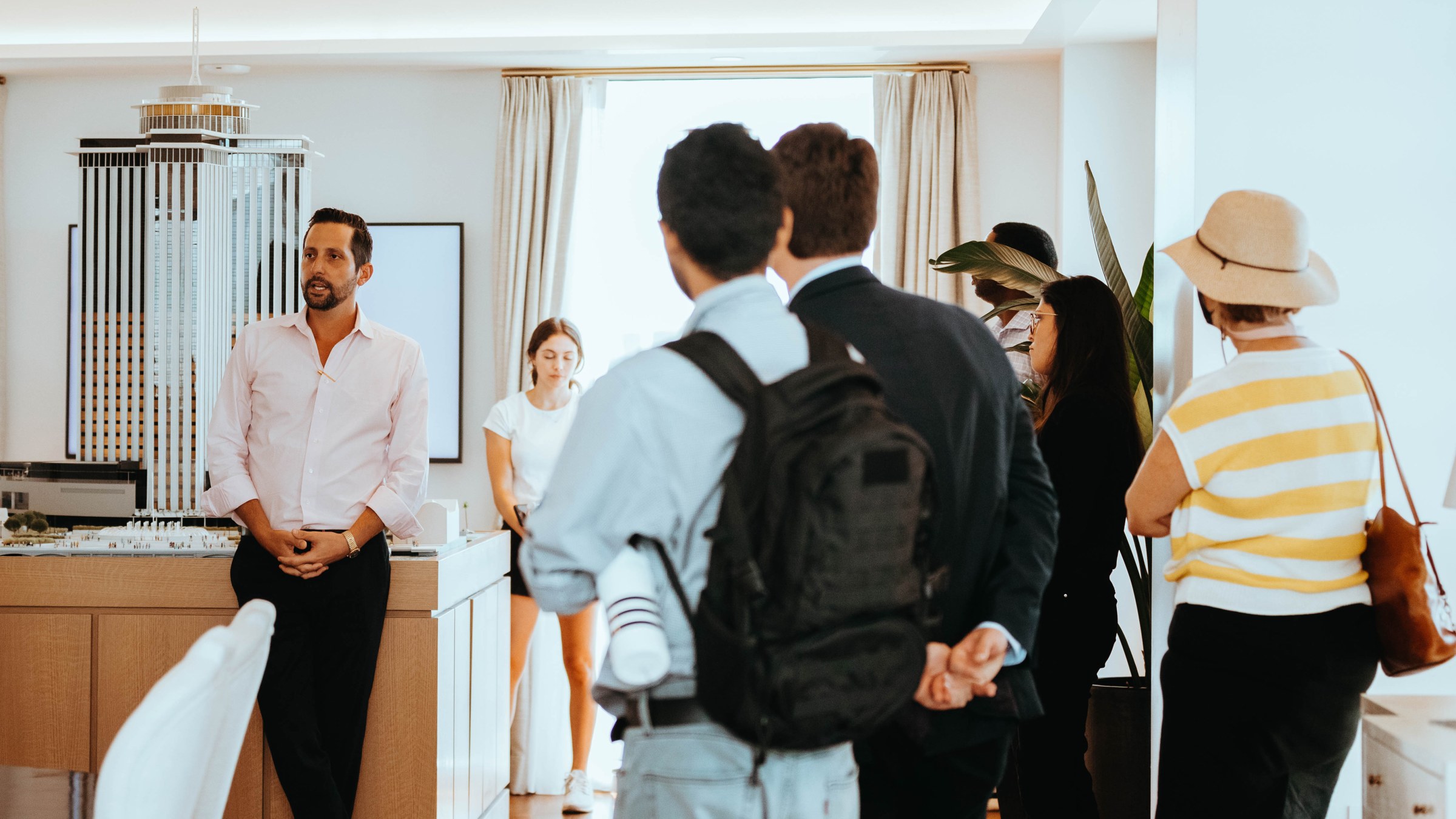 Students stand in a large room, backs facing the camera, while a real estate developer in the background left side speaks to them while standing in front of a large model of the redeveloped downtown New Orleans' Four Seasons building.