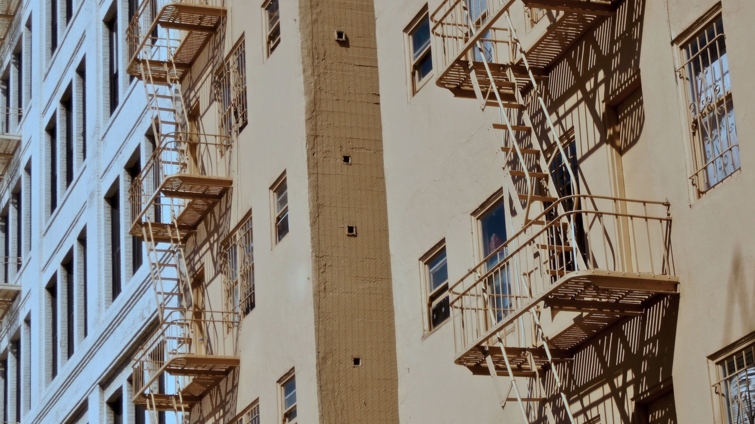 Side view looking up at three brick building facades with stacks of fire escape stairs and rows of windows.