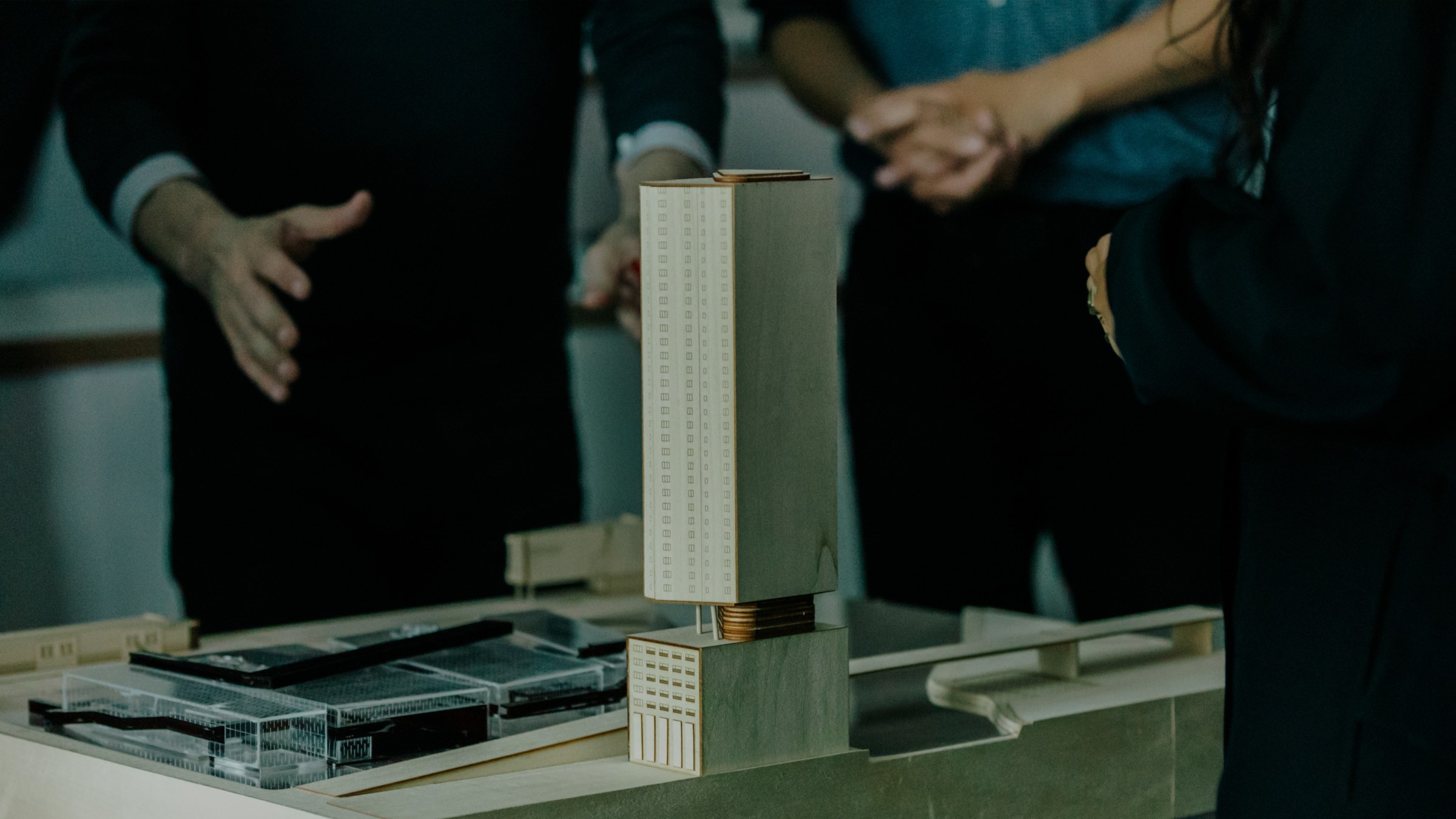 Close-up photo of a tall wooden tower and urban landscape model on a table with people standing behind it with only their torsos and hands in the frame.