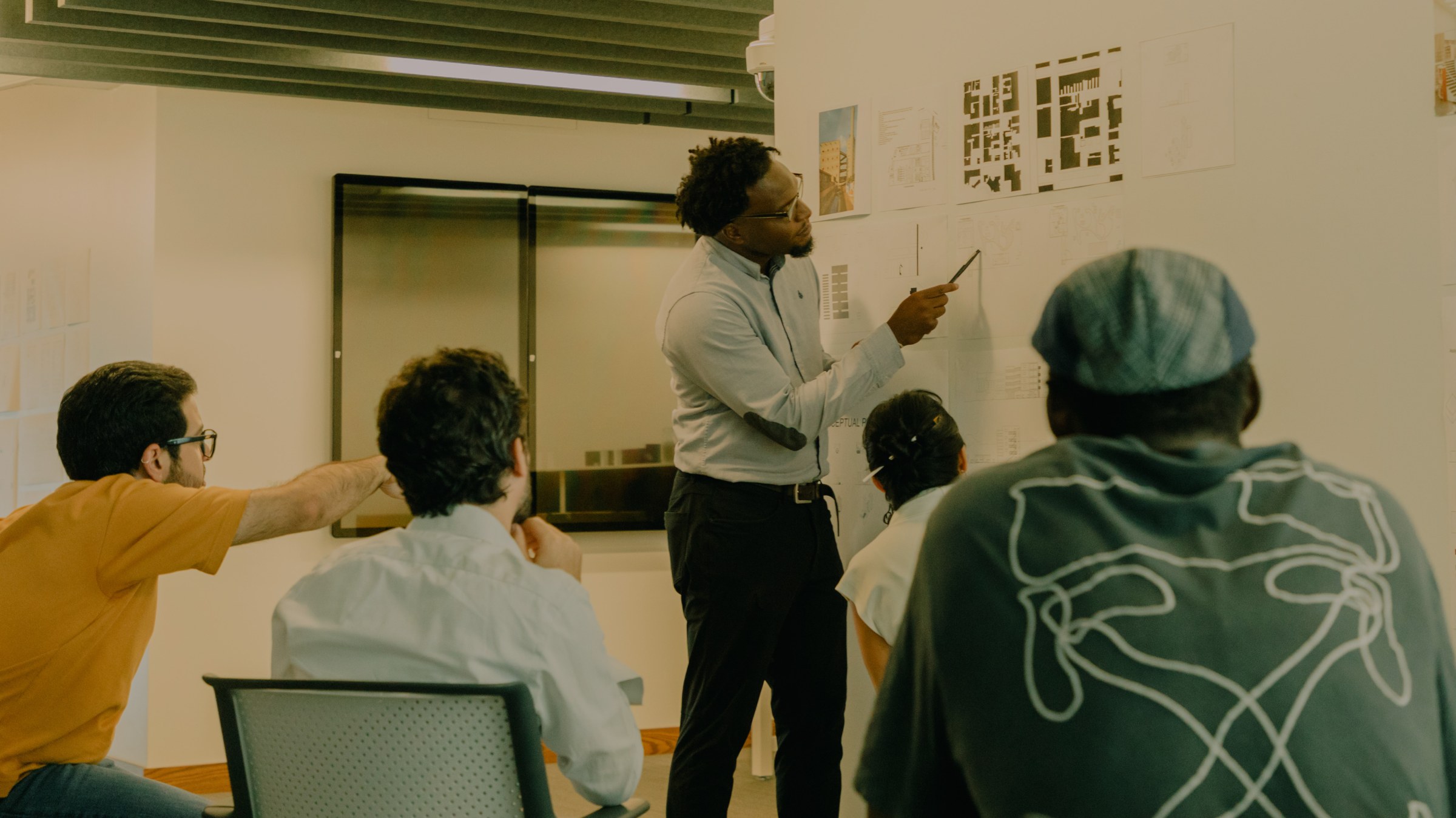 Student stands next to and points at pinned-up prints in the background while people sit in chairs, leaning forward, in the foreground with backs turned toward the camera's view.