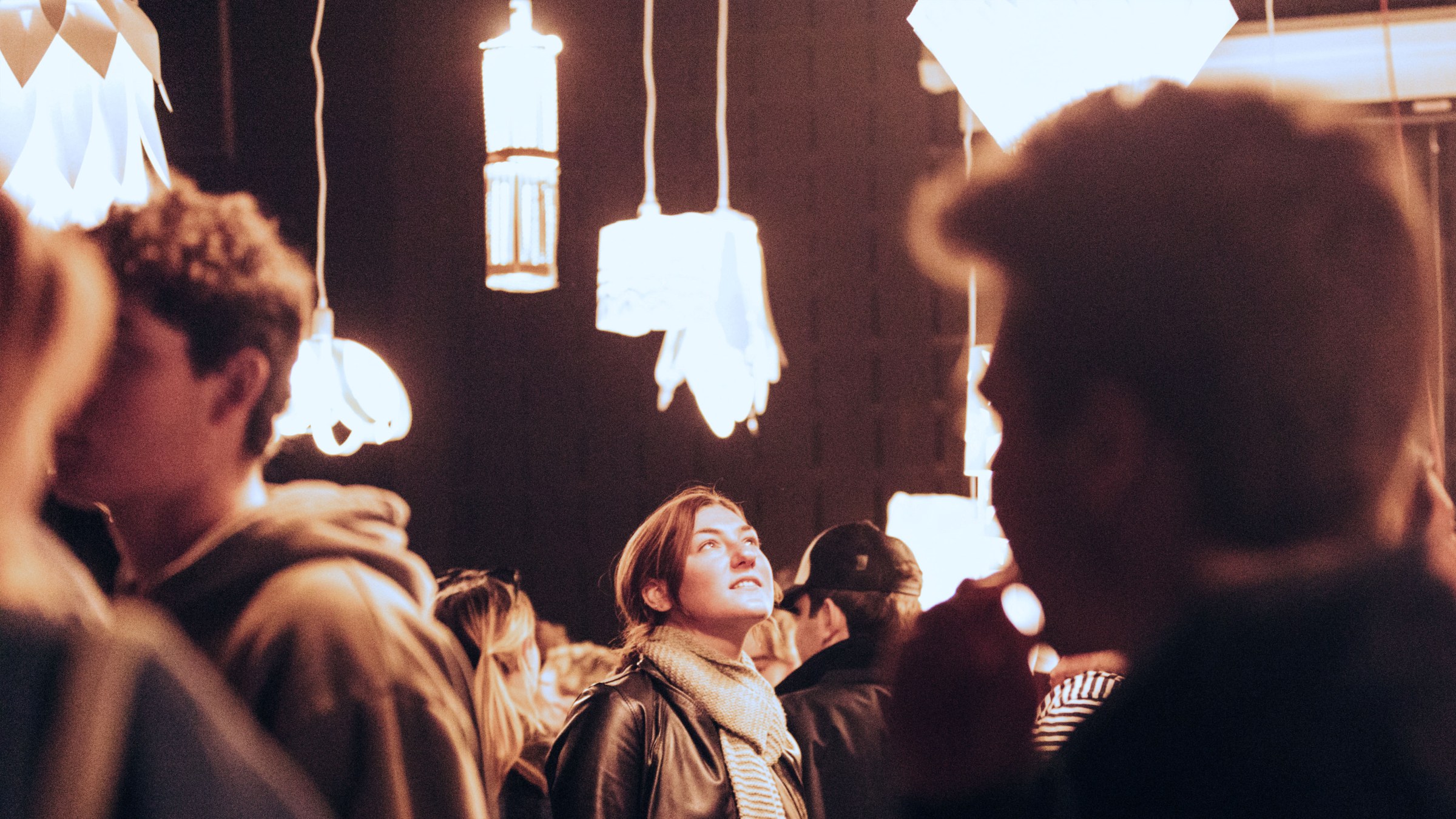 Light fixtures hang from the ceiling of a dark large crowded room, with one student looking up at a fixture in the center focus of the frame.