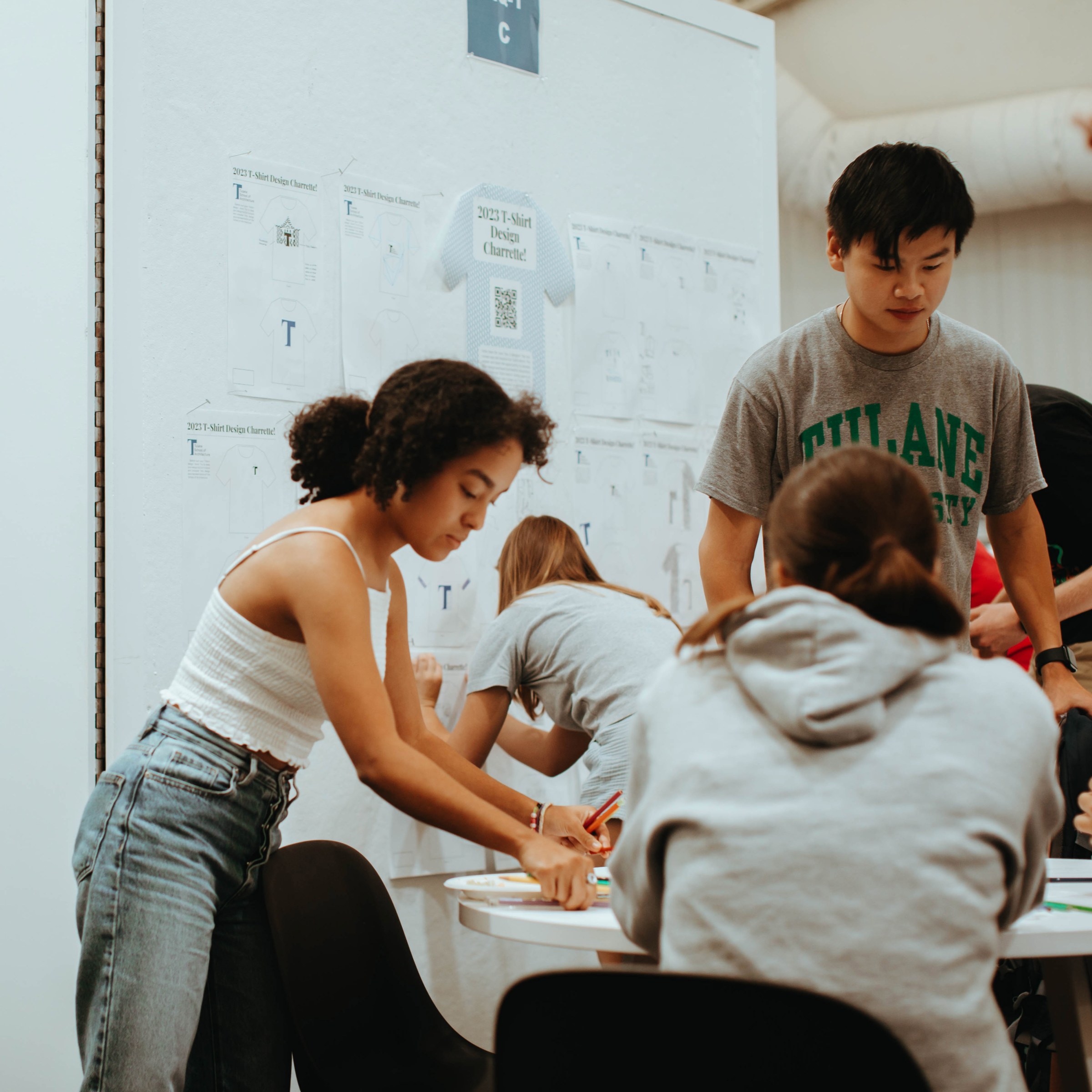 Four students sit and stand around a table working on a project with one of the students in the background, pinning up their drawing on a wall.