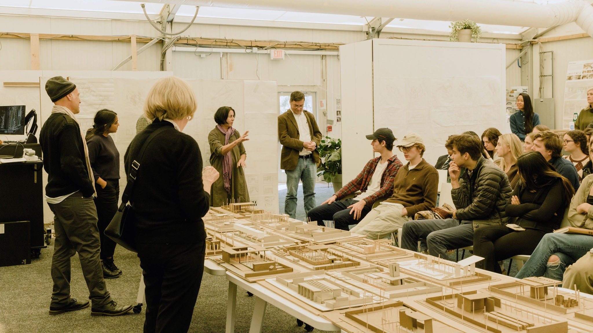 Students and faculty gathered together for a project review, study models sit on the table in front of them