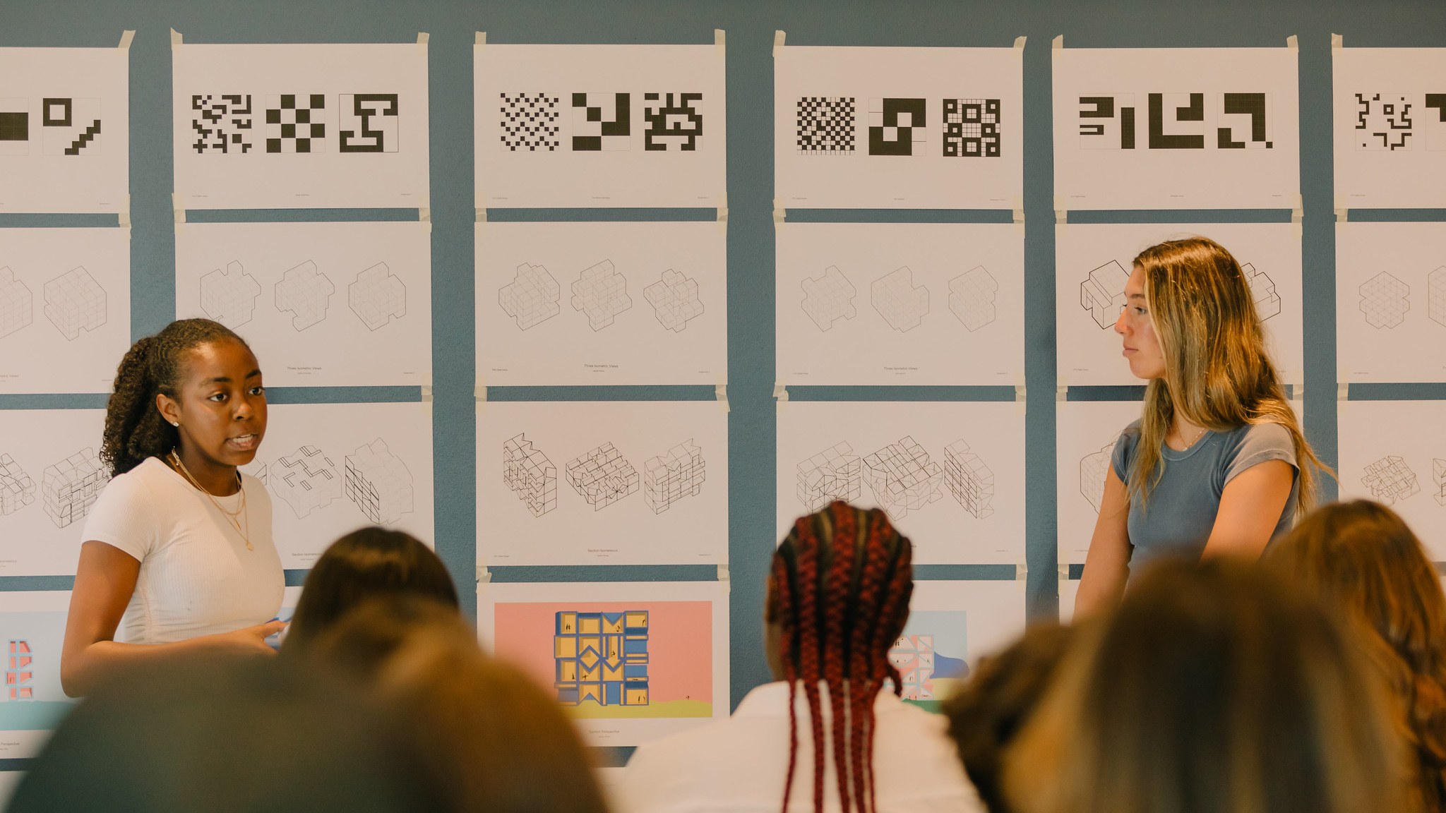 two students presenting their work to a group of people, their work is pinned up on the wall