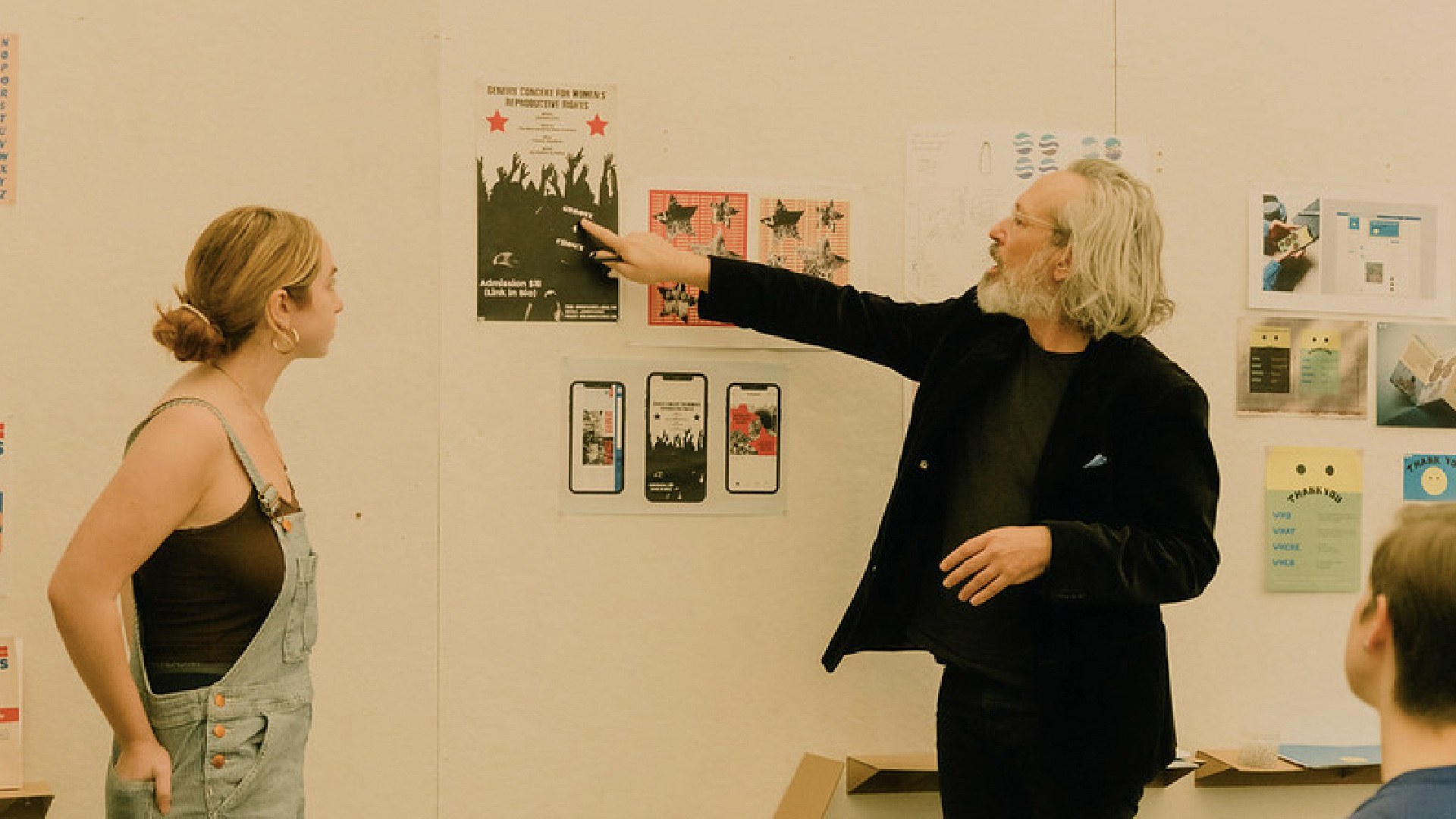 Image of a design class final review, with the student's work pinned up to the wall, while the student and the teacher discuss the work on the board.