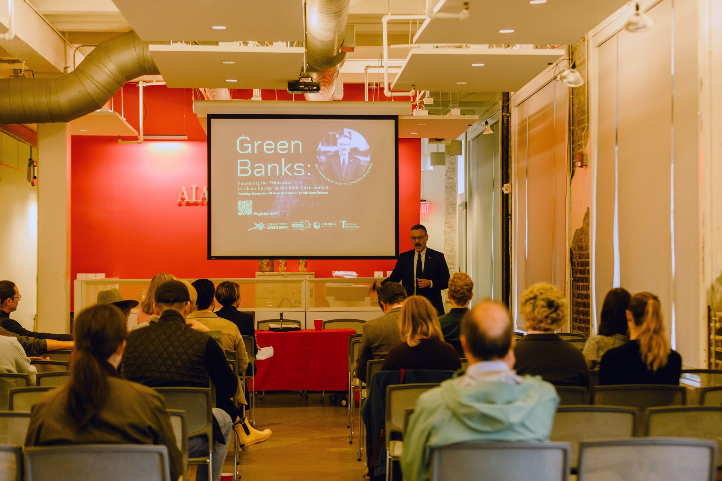 Photo of Connecticut Green Bank leader Bert Hunter standing at the front of a room with a projector presentation in the back ground and an audience with backs toward the camera in the foreground.