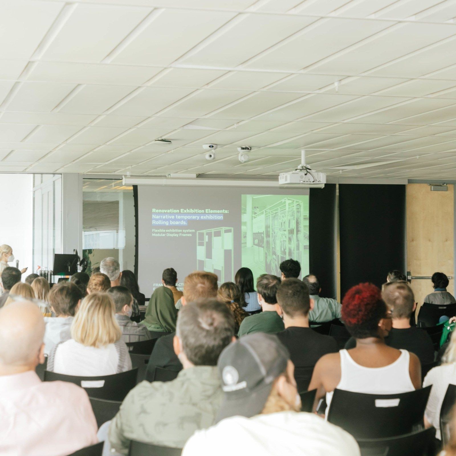 Photo from the back of a modern-design lecture hall, looking toward the front of the room, over the heads of many people sitting in chairs looking at slides on a projector screen.