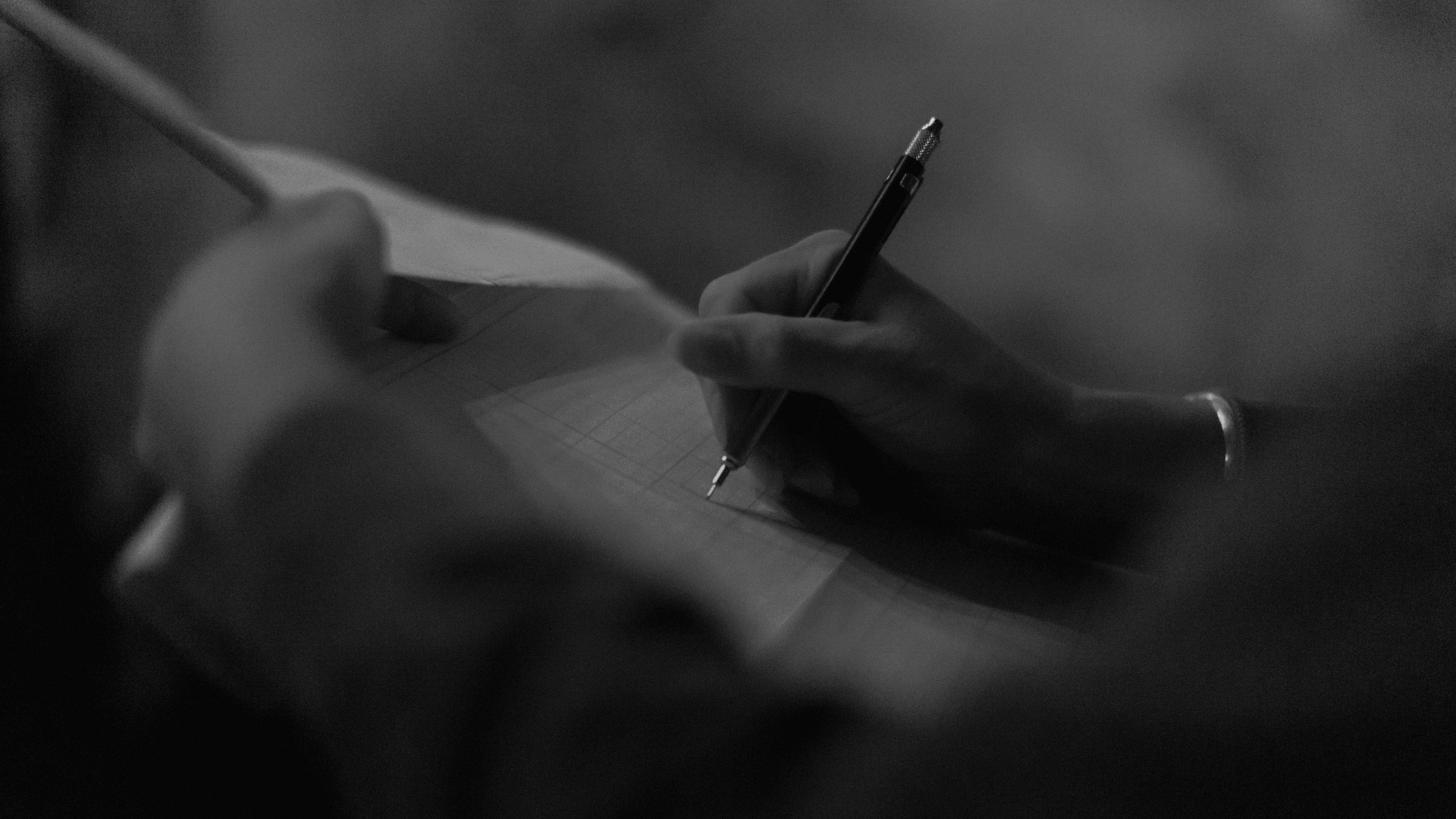 Close up of a hand holding a pen and writing on paper in a darkened room.