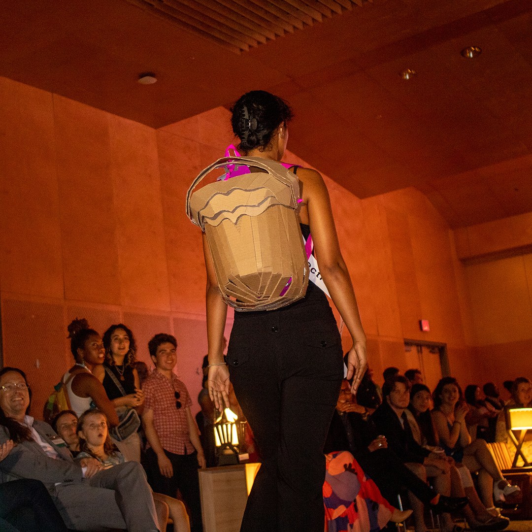 View of a model wearing a cardboard backpack on a runway inside a dark room with dozens of people sitting and standing in the background watching the fashion show.