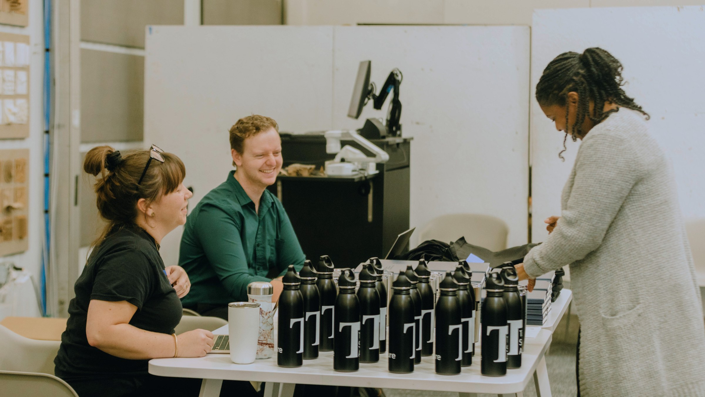 Two people seated on the left of a table topped with TuSA branded water bottles for sale, while a third person stands on the right of the table looking at the sale items.