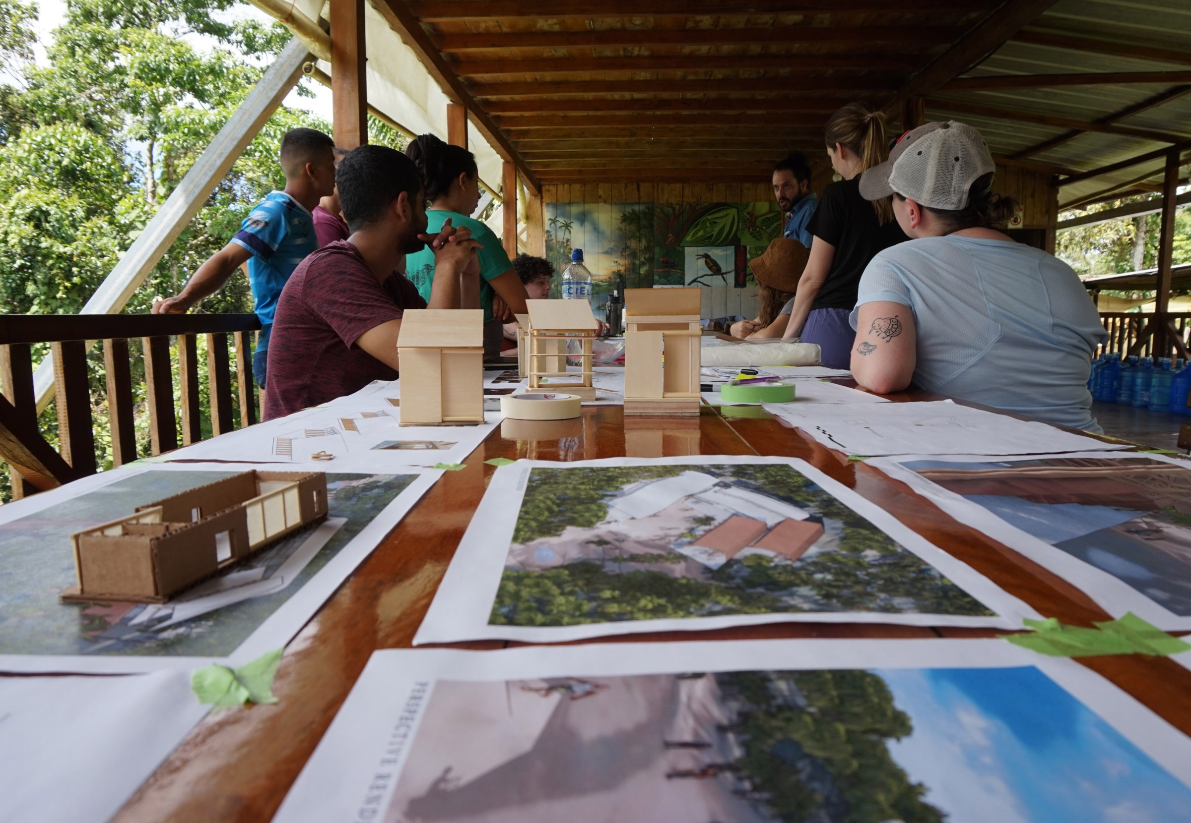 Architectural drawings and wood models cover a table in the foreground with people standing and sitting while talking in the background under a pavilion with a tropical forest on either side.