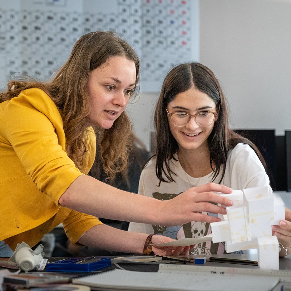 Faculty gives a student a desk critique of a 3D physical model, held by both faculty and student who are looking at the model, talking and student smiling.