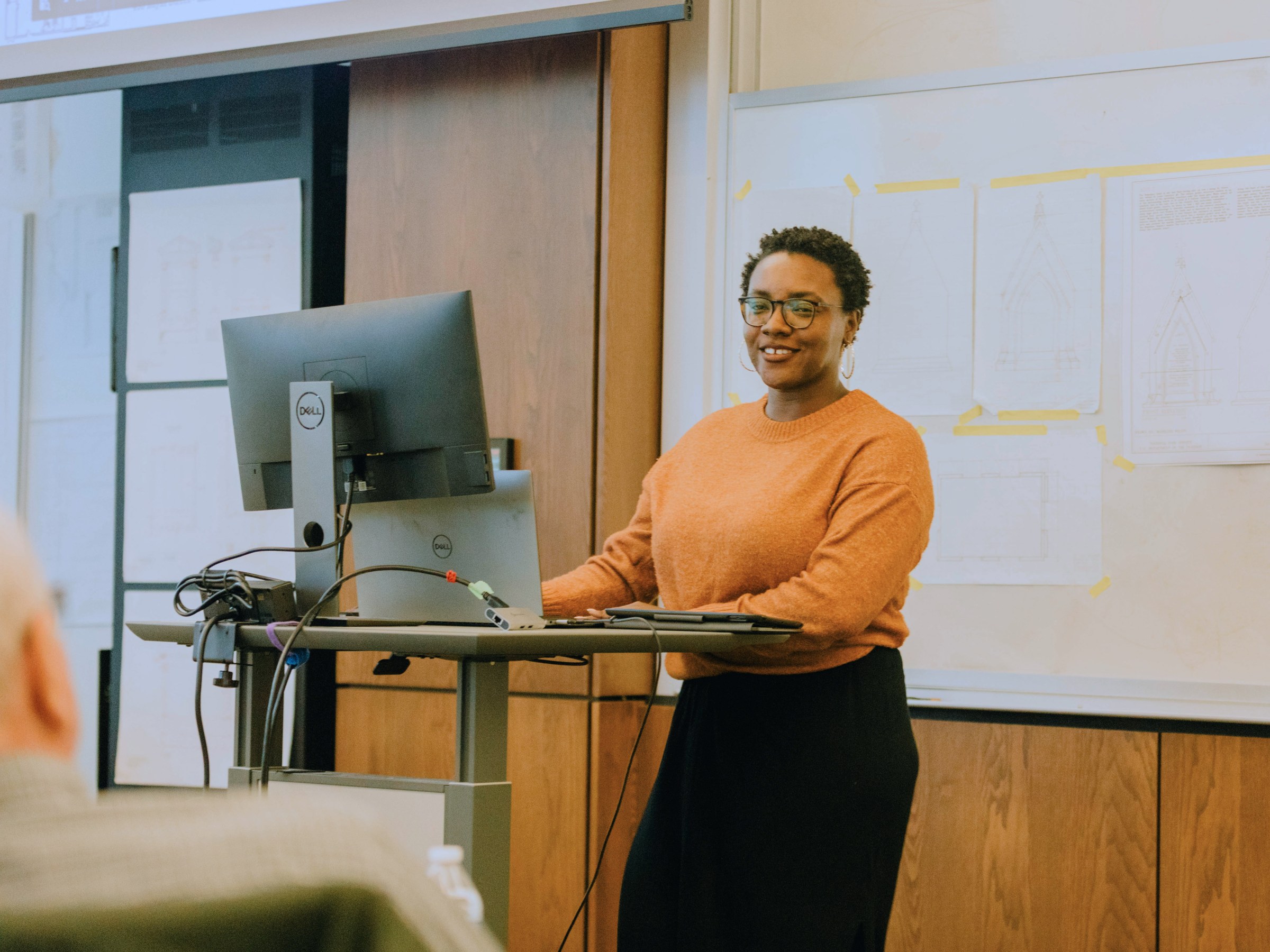 Tahlor Cleveland stands at a podium in the front of a classroom, smiling while giving a presentation for a studio.