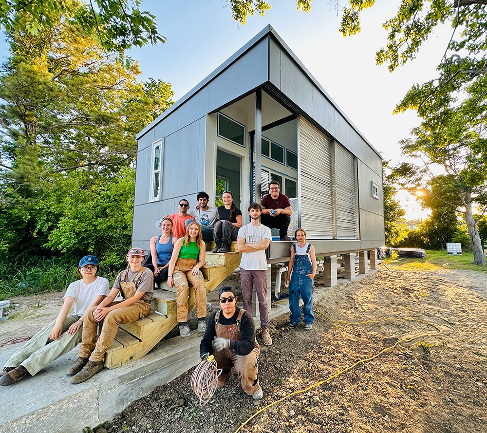 Group photo of students sitting and standing in front of a tiny house built for a homeless man lots of trees around the edges of the home.
