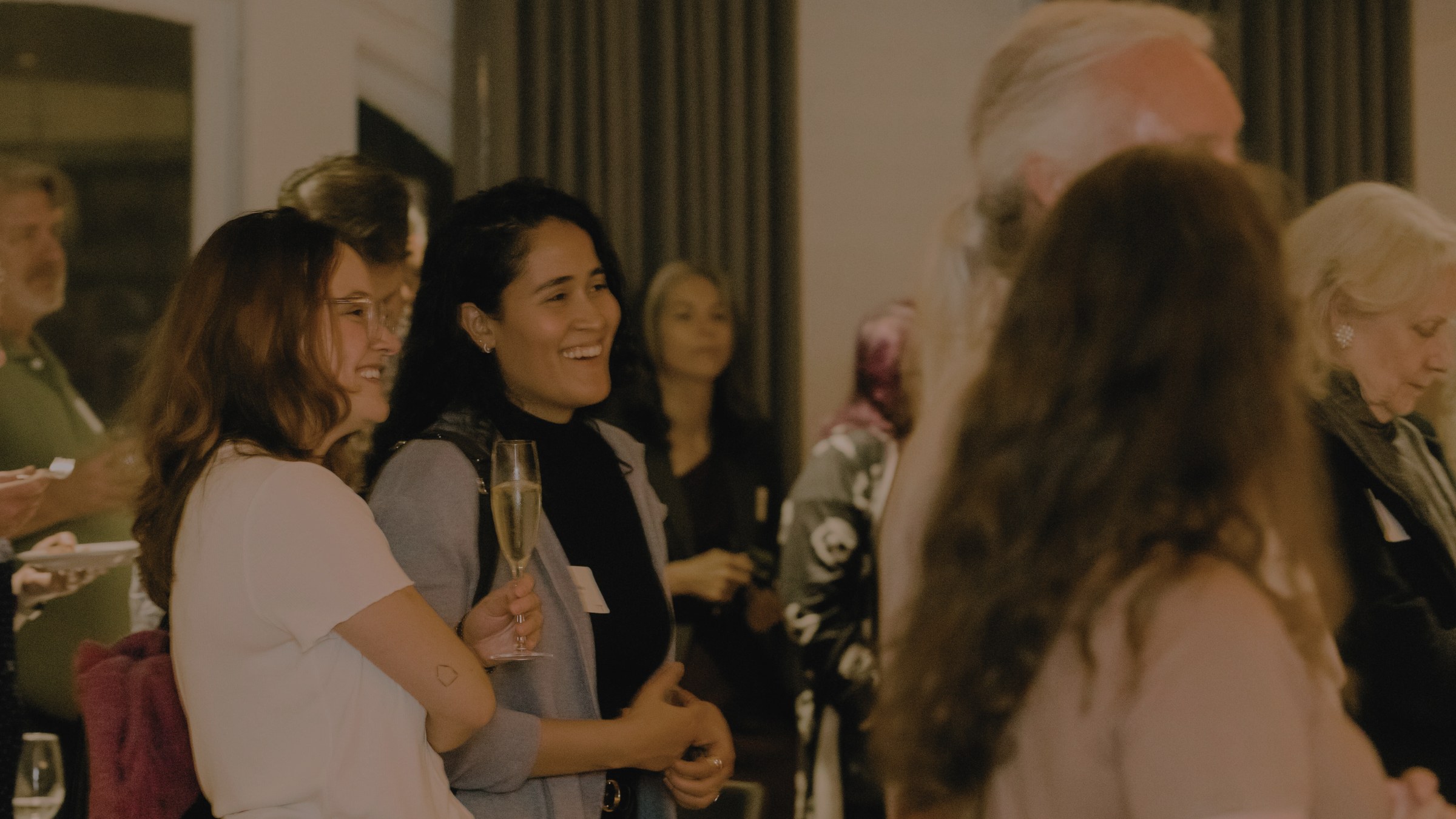 Photo of two women laughing while standing amongst a crowd at an alumni reception.