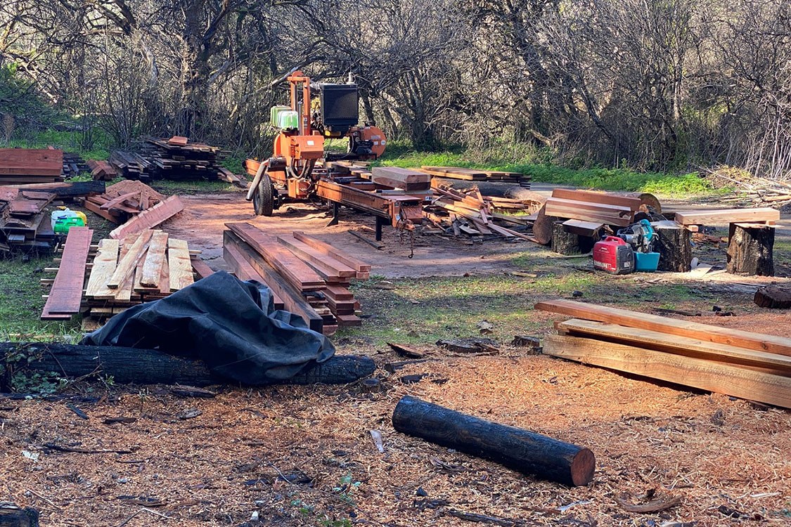 Lumber yard with stacks of wood and other materials in the foreground and a cutting machine in the background.