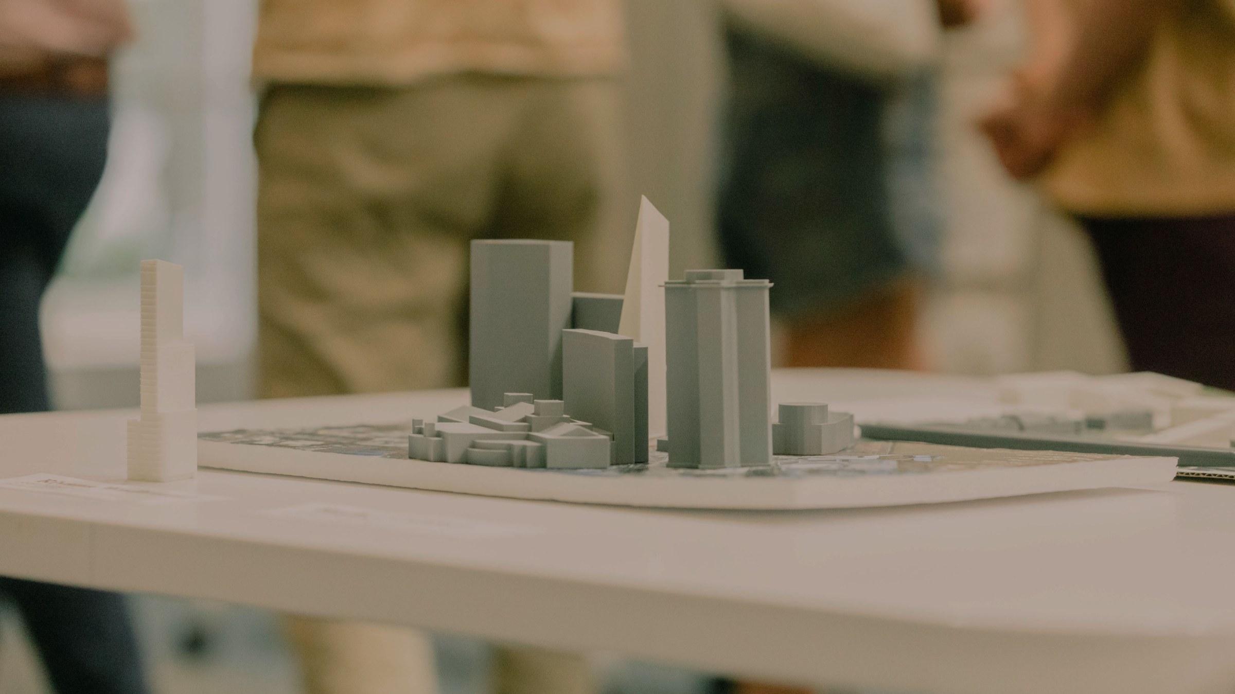 close-up photo of a site model of an urban setting with multiple tall buildings, standing alone on a table with a few people standing in the far background.