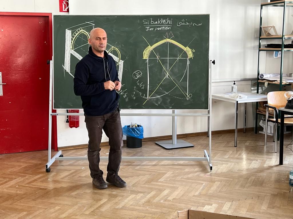 Eisa Esfanjary stands in front of a chalkboard at the front of a room.