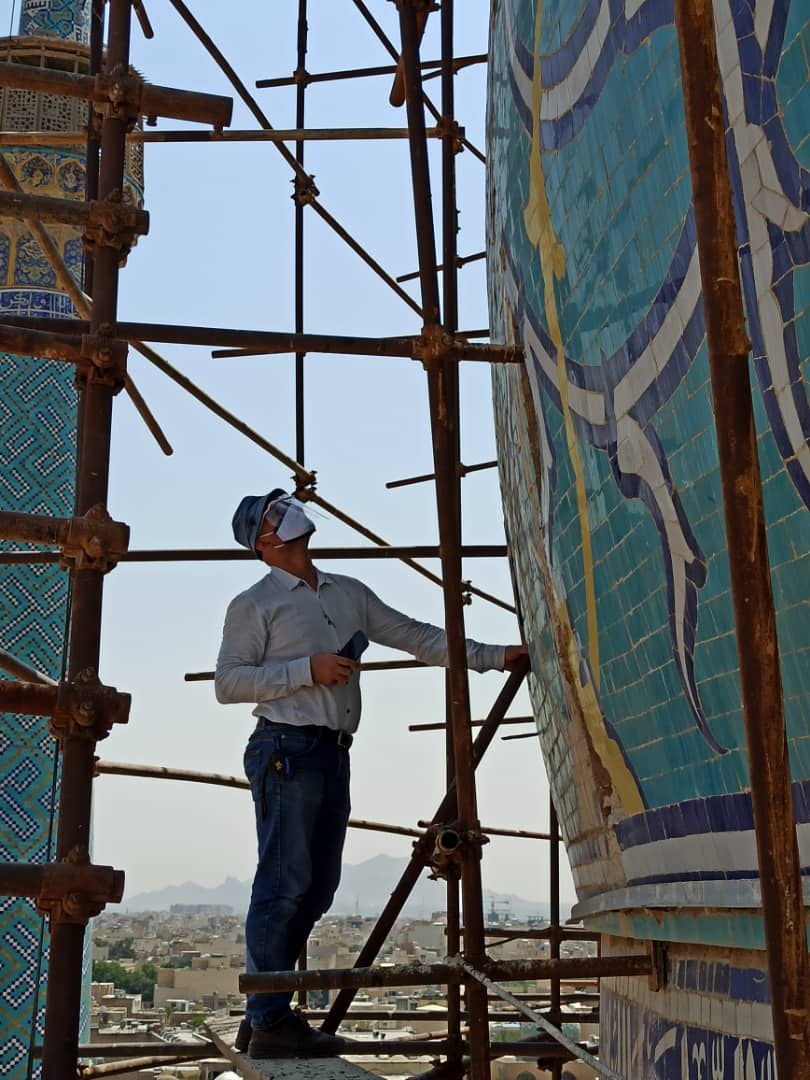 Eisa Esfanjary stands on scaffolding, looking up at a tile mosaic wall outside, overlooking an urban skyline in the background.