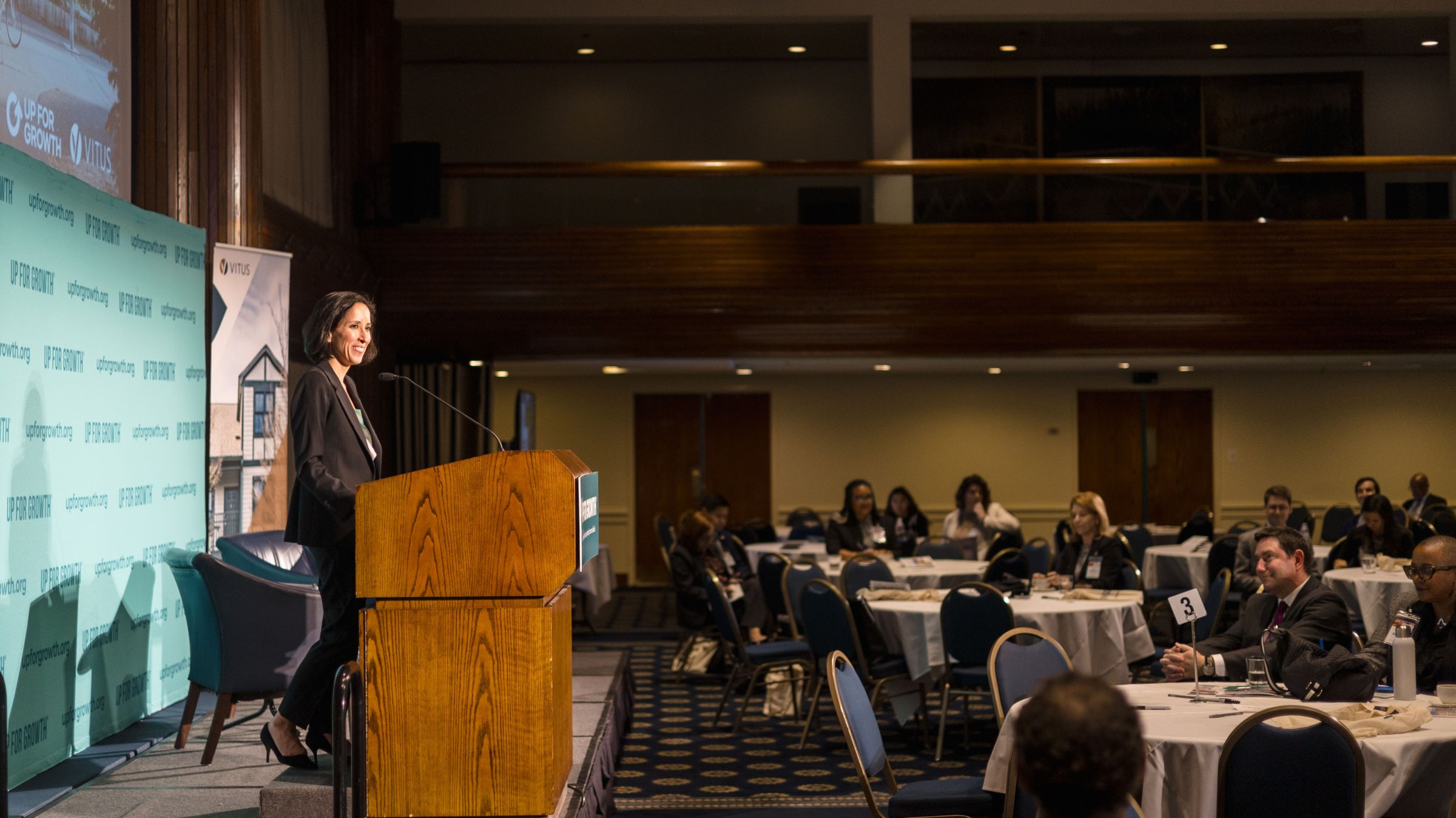 Side view of Sara Bronin speaking at a podium on a stage in a large conference hall with the audience seated at roundtables to the right.